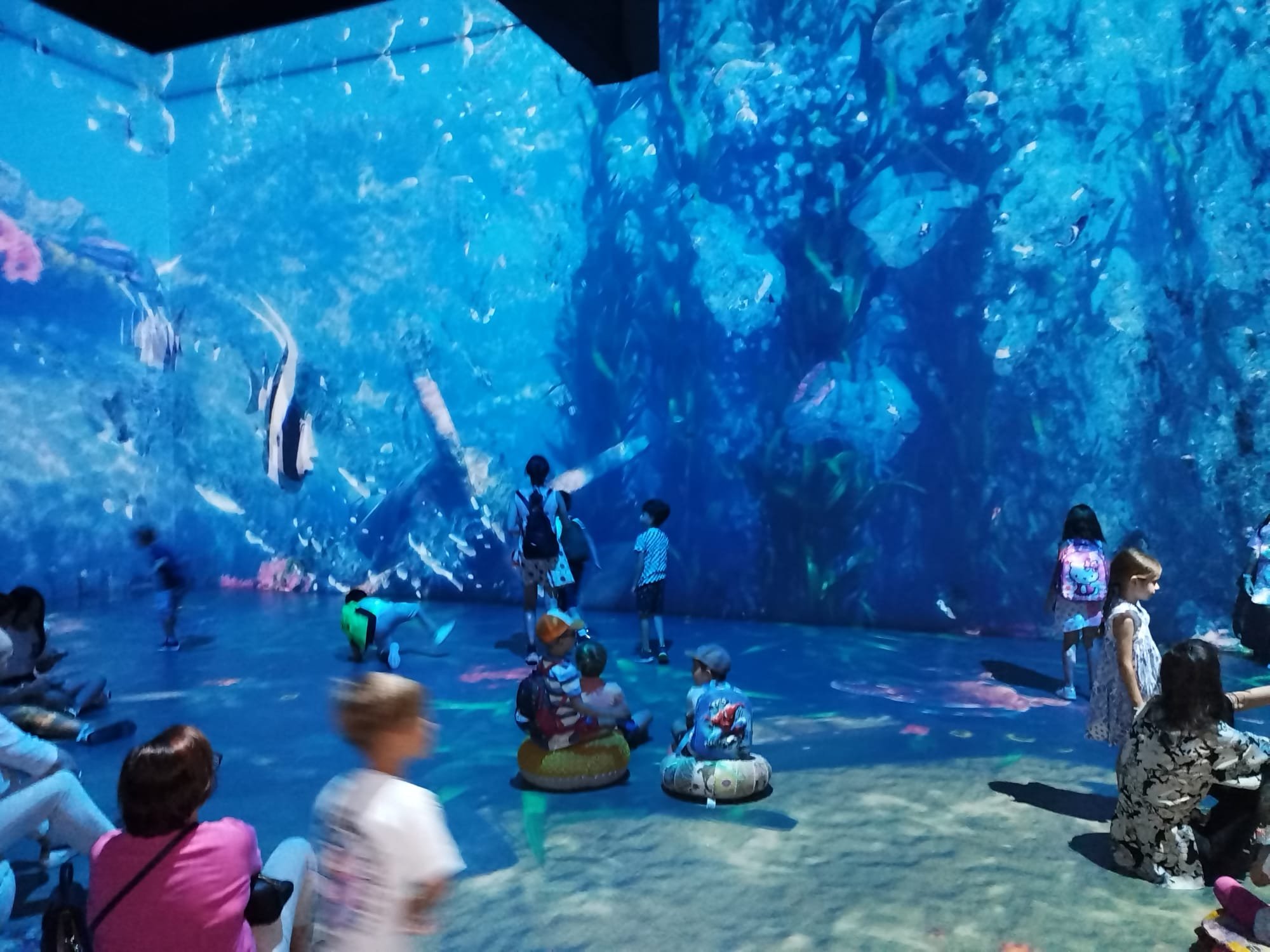 Children and adults engaging with an underwater-themed digital projection on walls, featuring fish and ocean visuals, at an interactive exhibit.