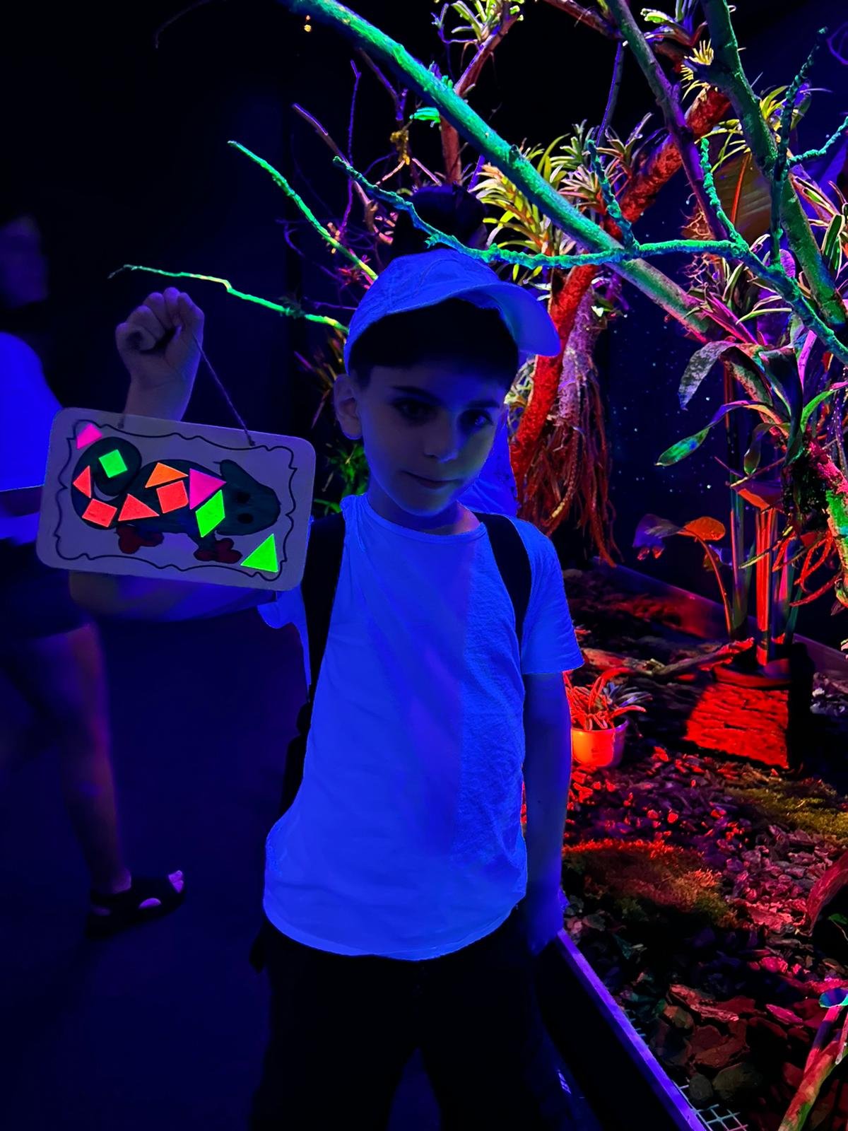 A young boy in a white T-shirt and cap holding a glowing craft under black light, standing in a neon-lit exhibit with plants and vibrant colors.