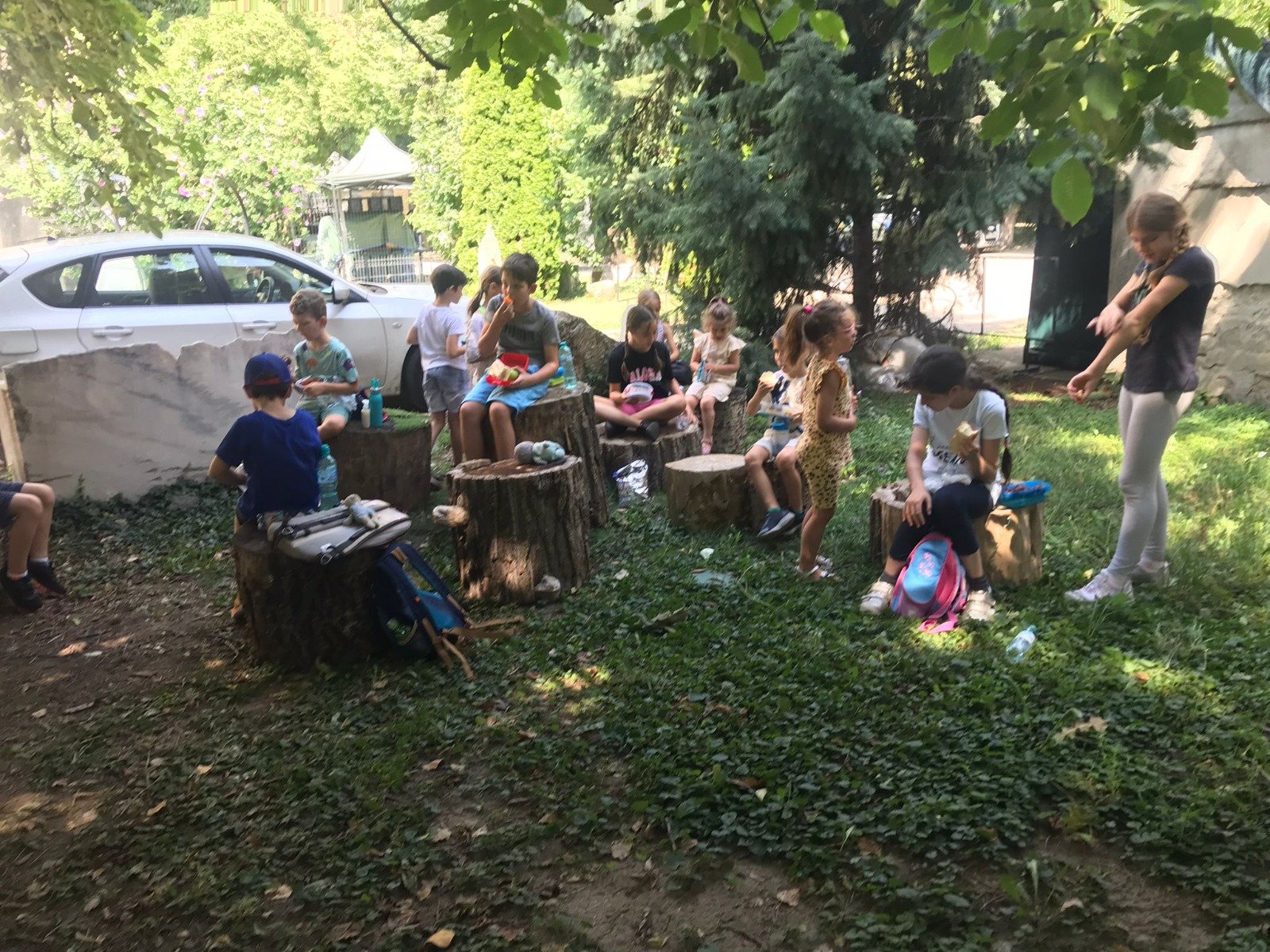 Children sitting outdoors on tree stumps, having a picnic or lunch, with a white car and greenery in the background.