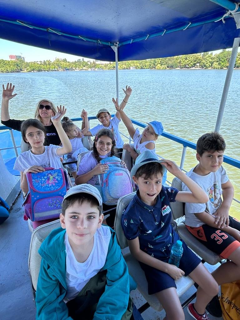 Group of children and an adult on a boat, wearing casual summer clothes and hats, holding backpacks, with a lake and trees in the background.