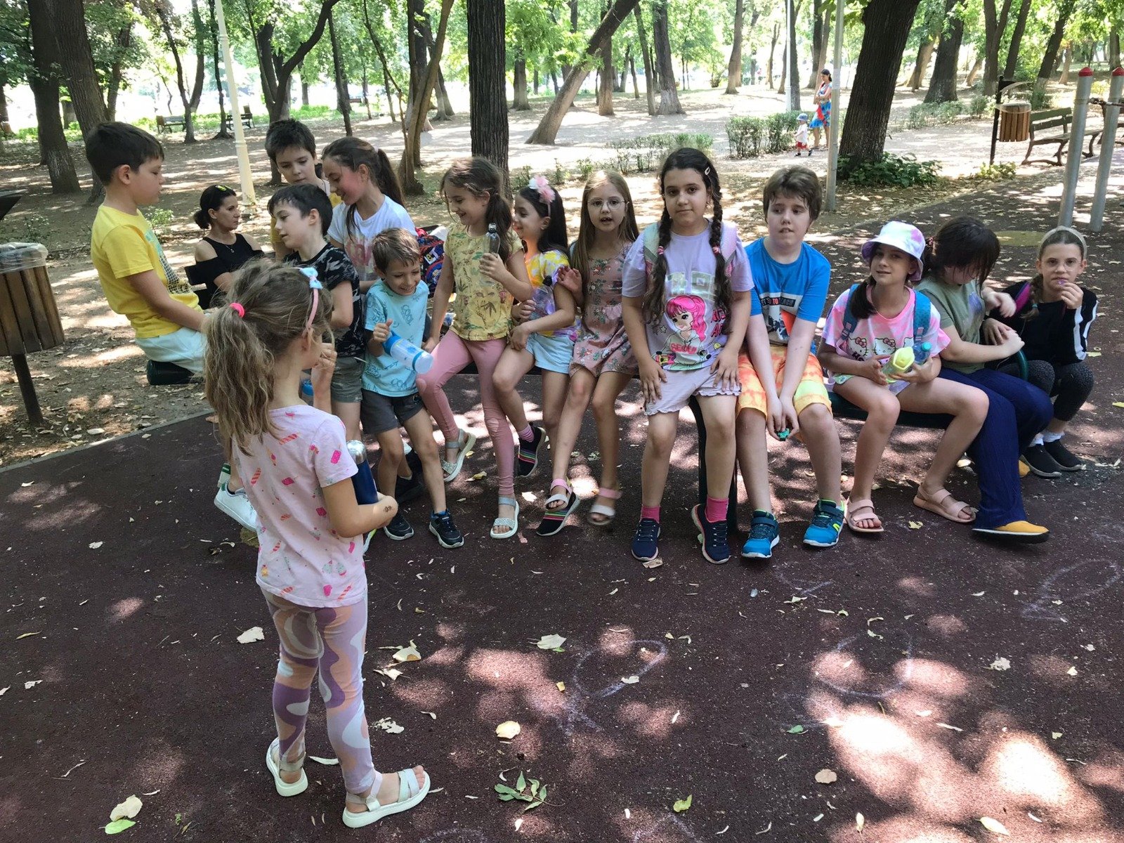 Group of children sitting and standing in a park, surrounded by trees, on a sunny day.
