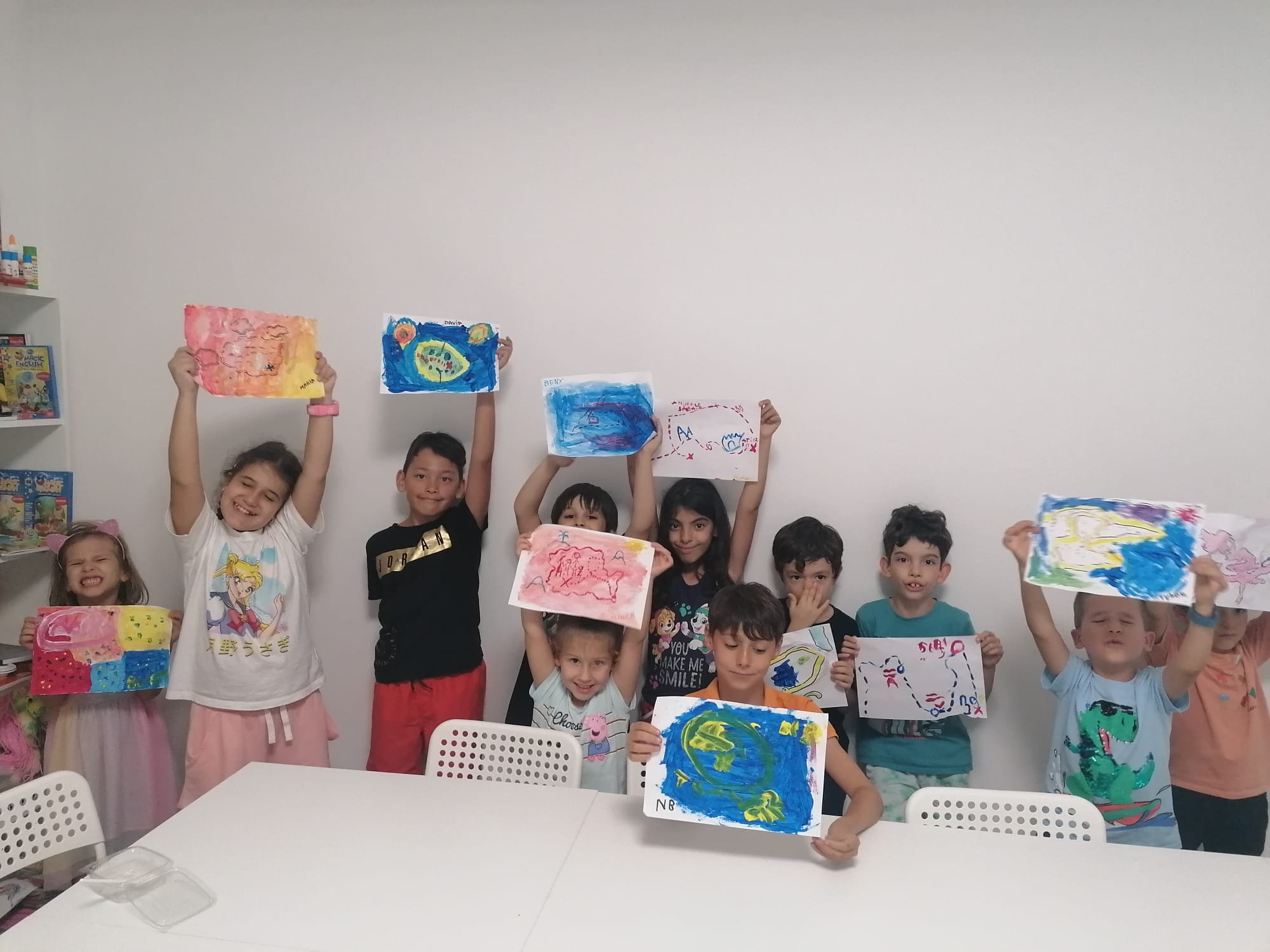 Group of children holding colorful artwork, standing indoors, white table in foreground, shelves with art supplies in background.