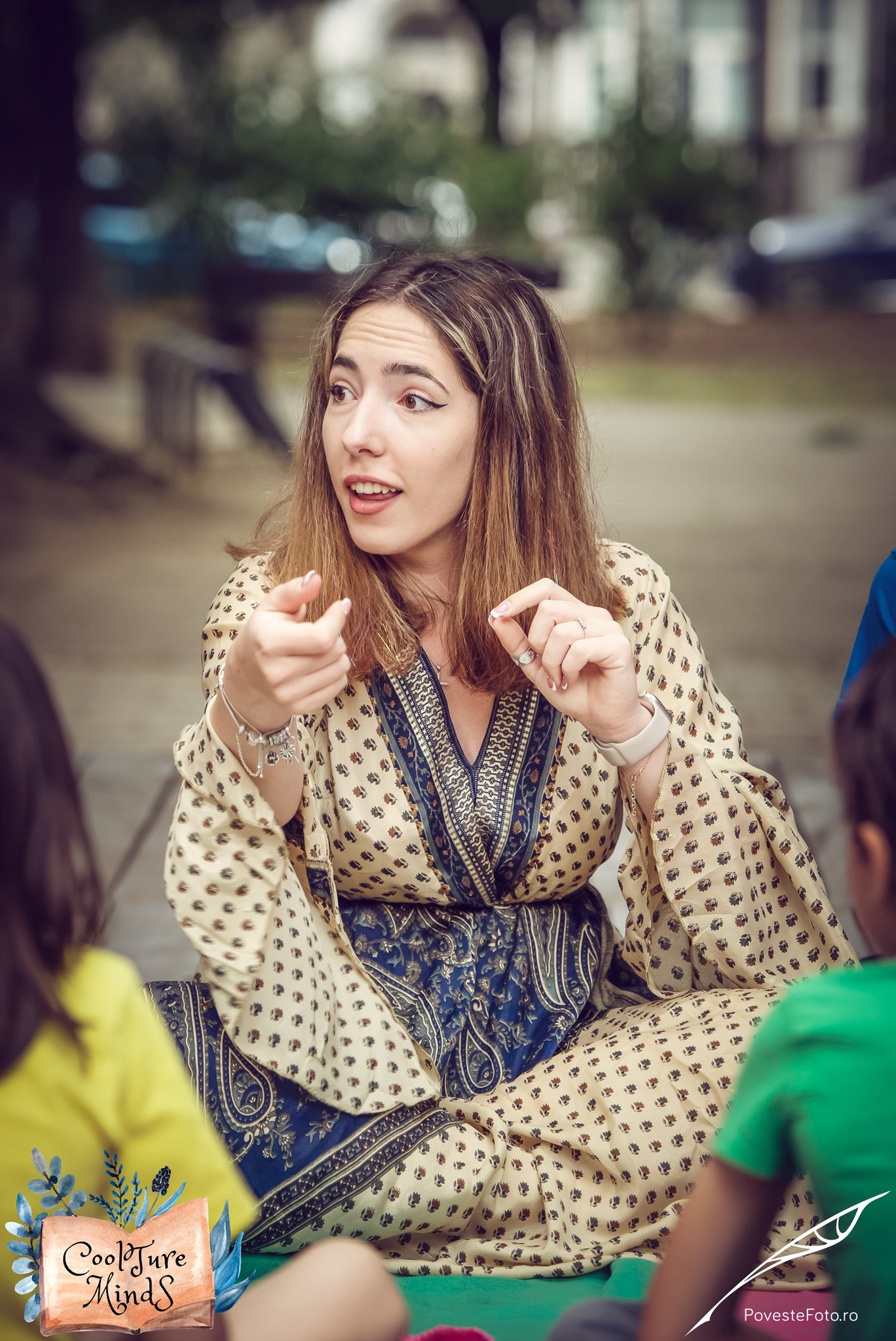 Woman in a patterned dress speaking to children outdoors, seated on a colorful blanket, with a "Coolture Minds" and "PovesteFoto.ro" logo on the image.