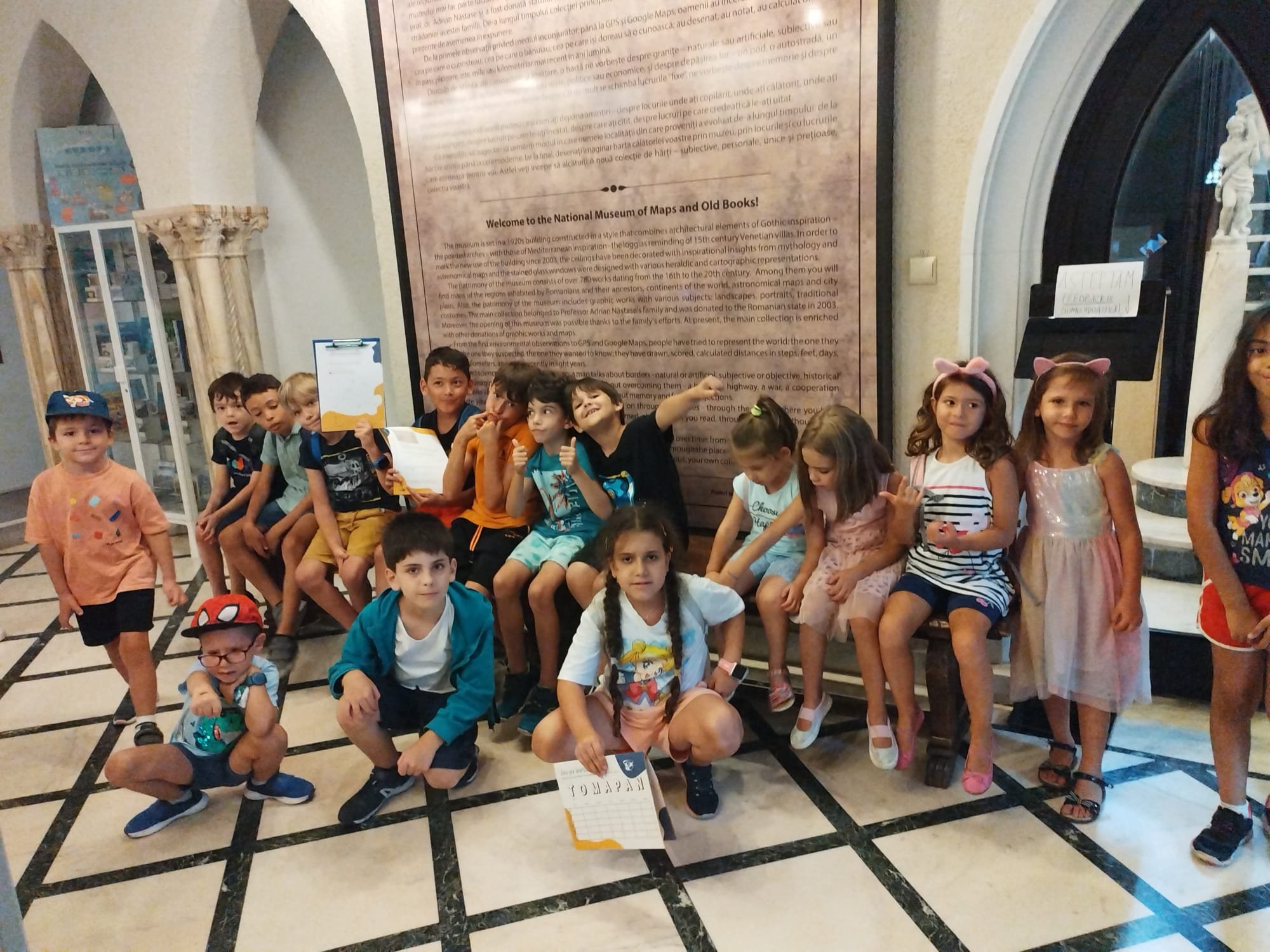 A group of children sitting and standing indoors, some holding signs, in front of a display labeled 'Welcome to the National Museum of Maps and Old Books.' One child is wearing a hat with ears and several are dressed casually. The room has a tiled fl