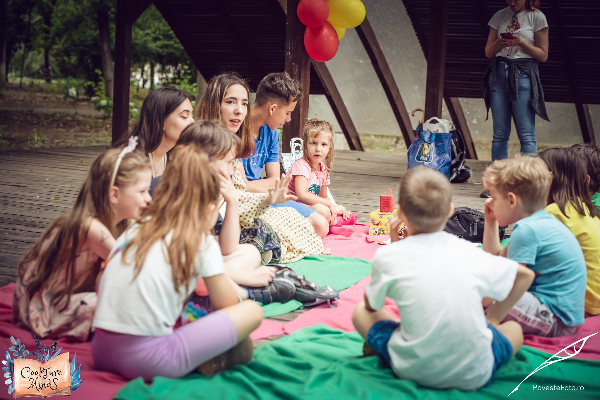 A group of children and a young woman sitting on colored blankets for an outdoor activity, under a wooden structure with balloons. Some children are engaged in conversation, while another person stands in the background using a phone. There are bags