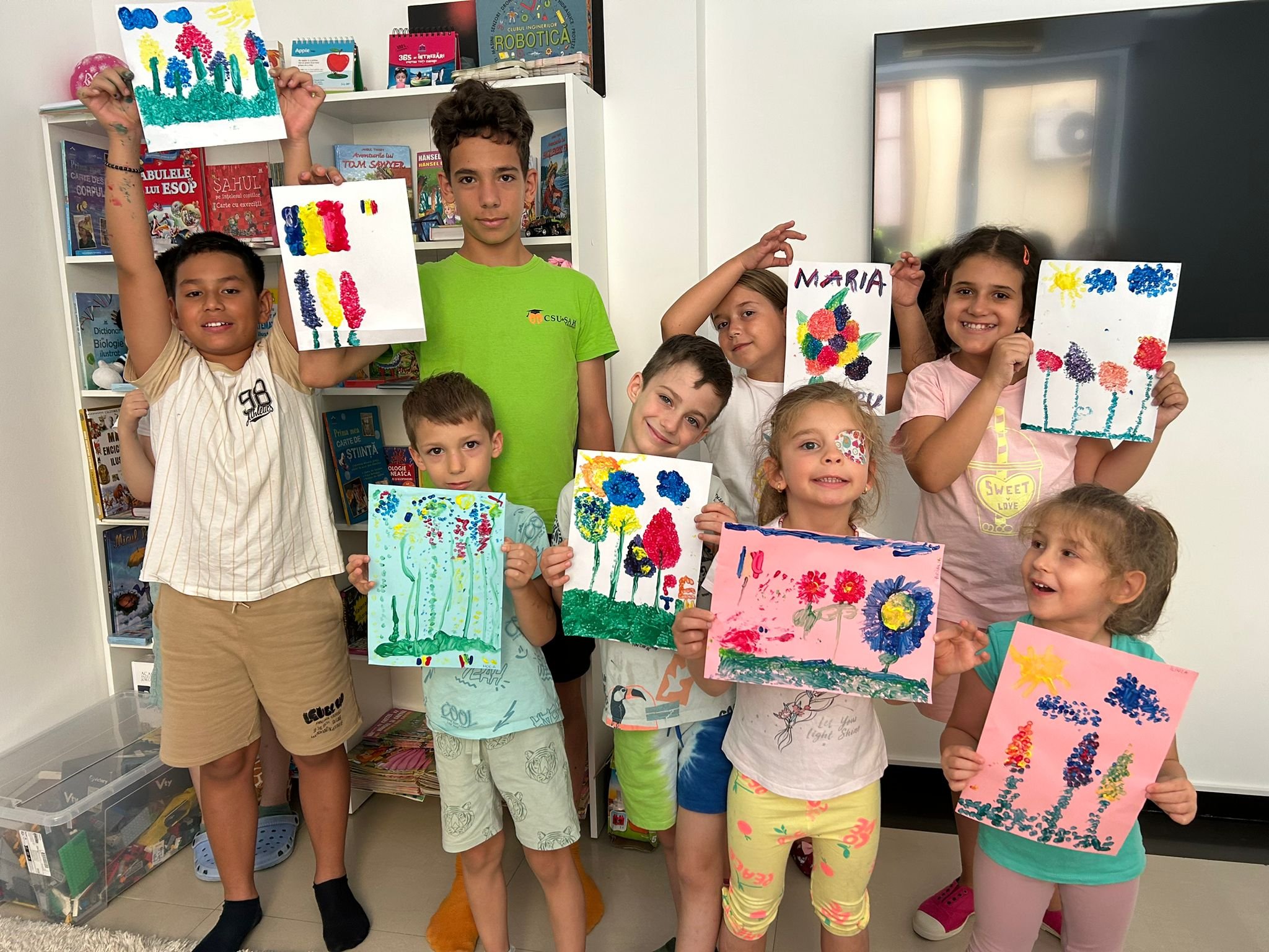 A group of children holding colorful paintings of flowers in a classroom setting, with bookshelves in the background.