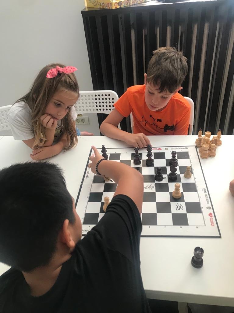 Three children playing chess at a white table, with one child making a move while the other two watch attentively.