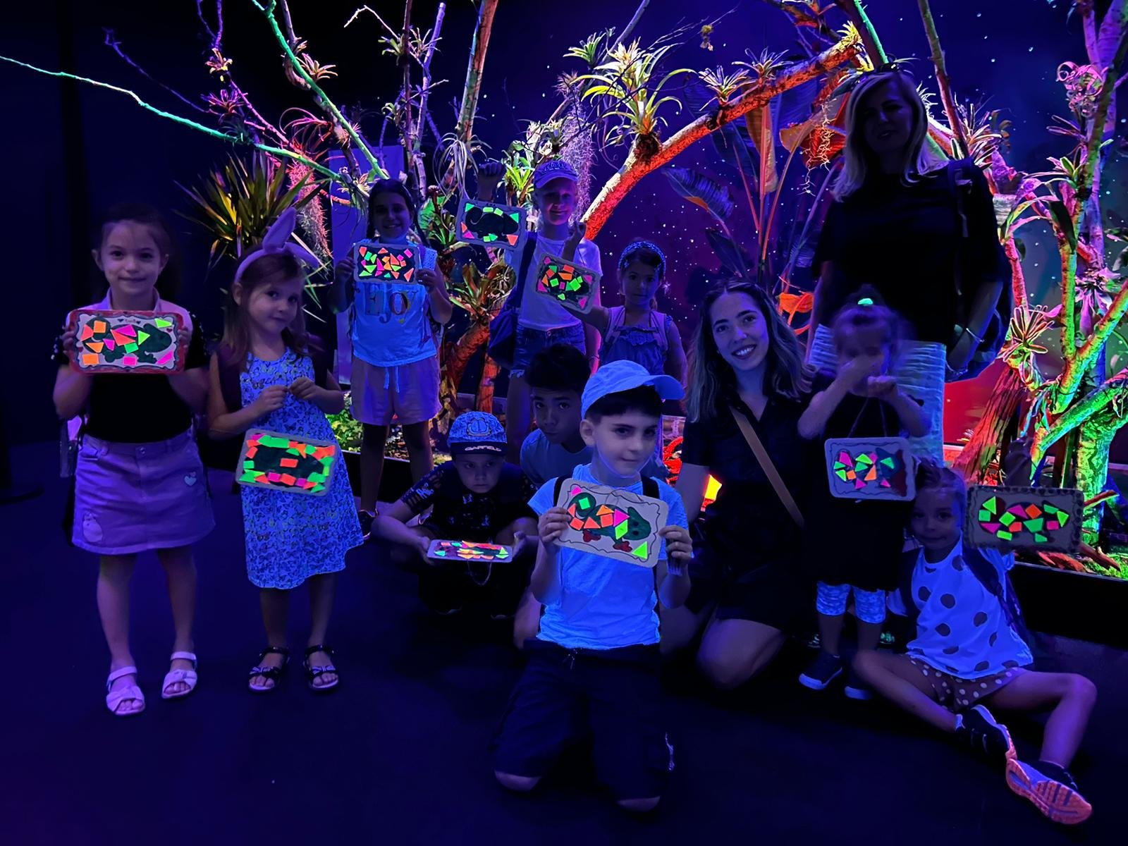 Children and adults in a neon-lit room holding colorful artworks made with glow-in-the-dark materials.