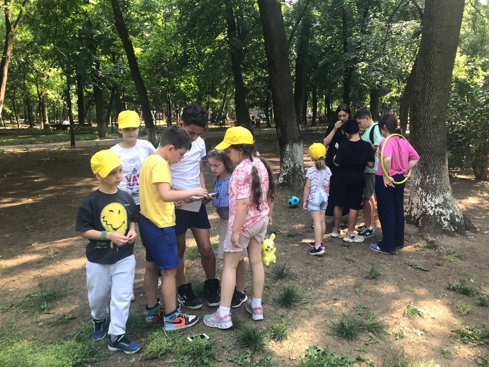 Group of children wearing yellow caps and casual clothes standing and sitting in a park with trees and grass.