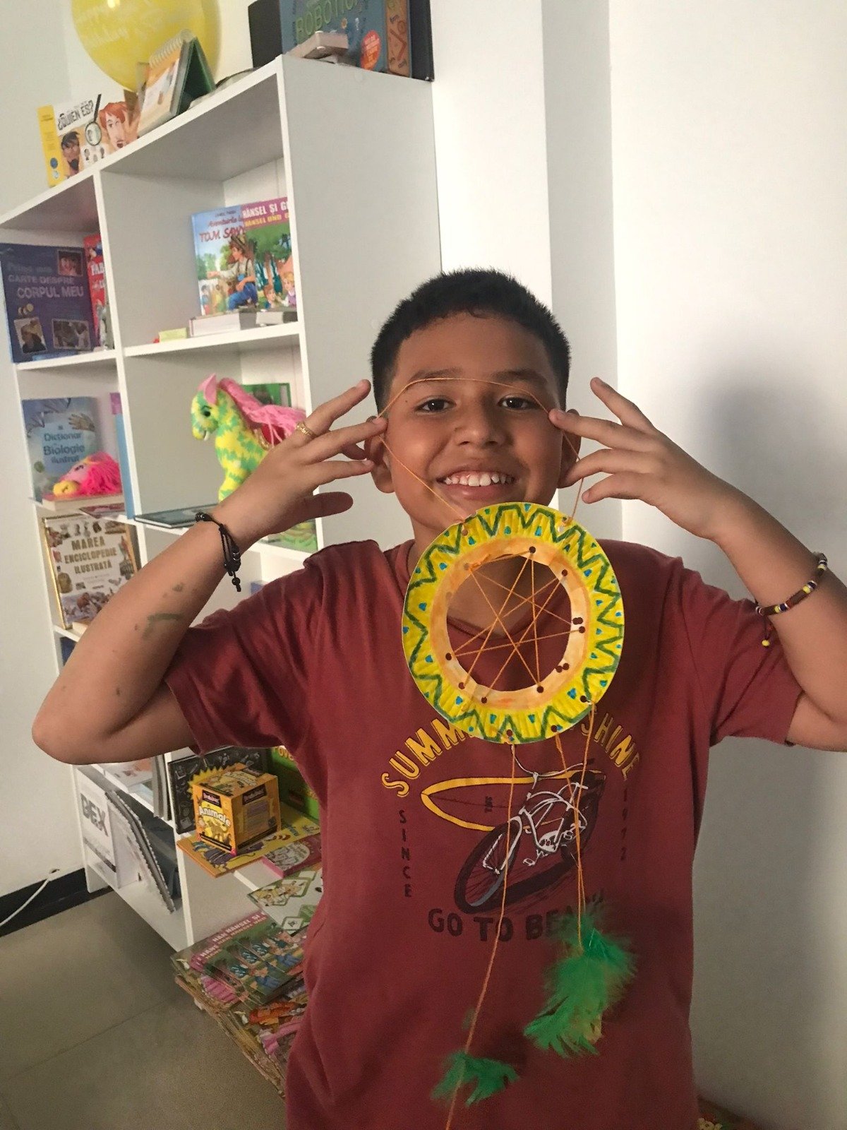 Smiling child holding a colorful handmade paper dreamcatcher in front of a bookshelf with toys and books in the background.