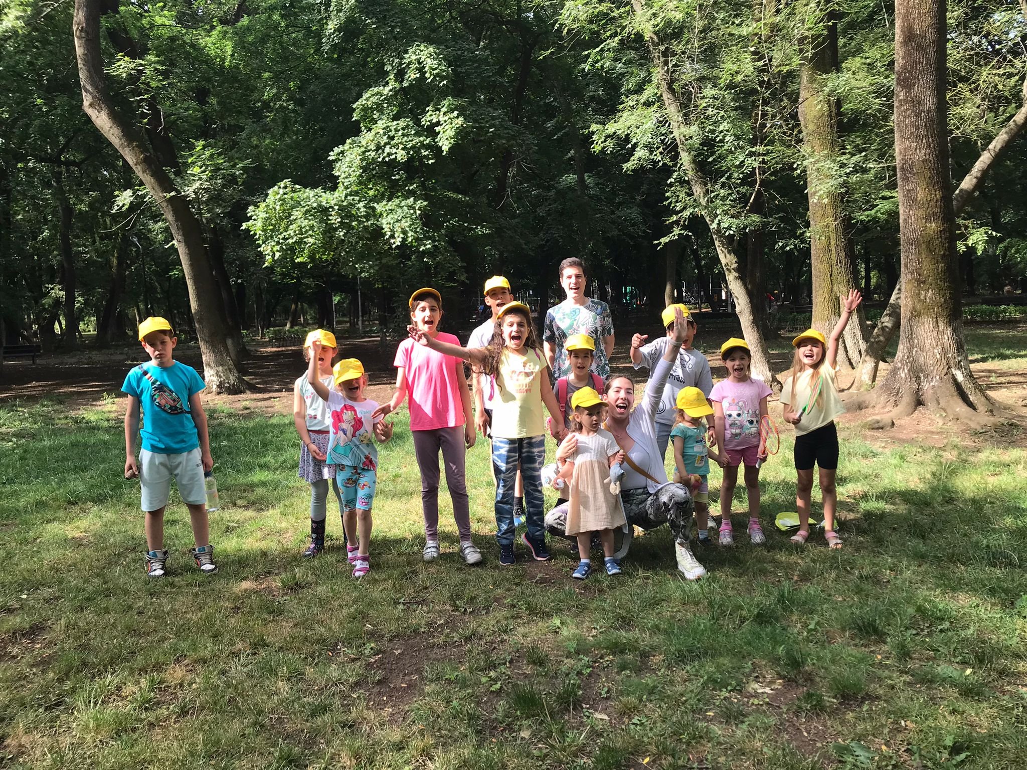 Group of children and adults wearing yellow hats posing in a park with trees and grass.