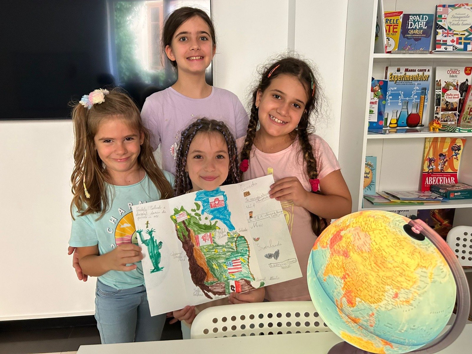 Four young girls holding a colorful drawing, with a globe and books in the background.