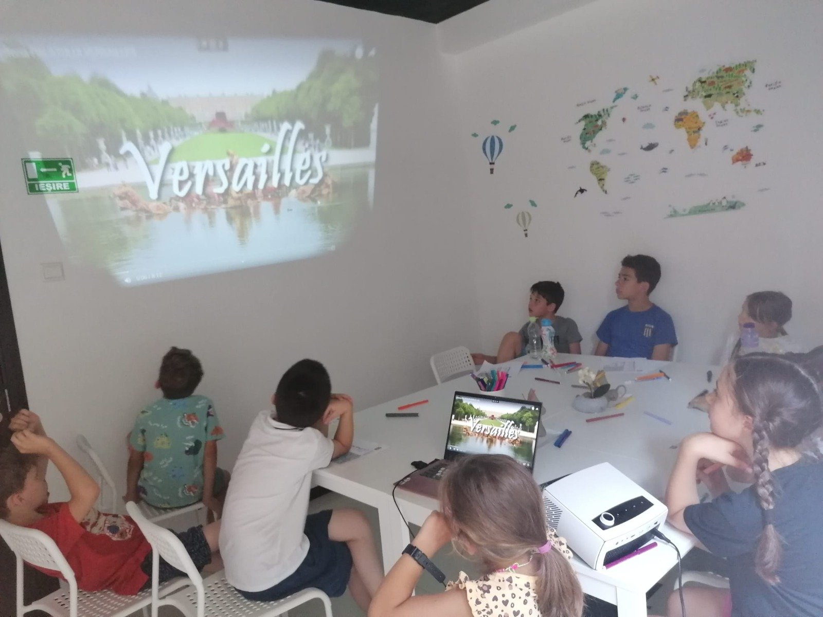 Children watching a projection of "Versailles" on a wall in a classroom setting, with a laptop and projector on a table, surrounded by school supplies, with a world map and decorative stickers on the wall.