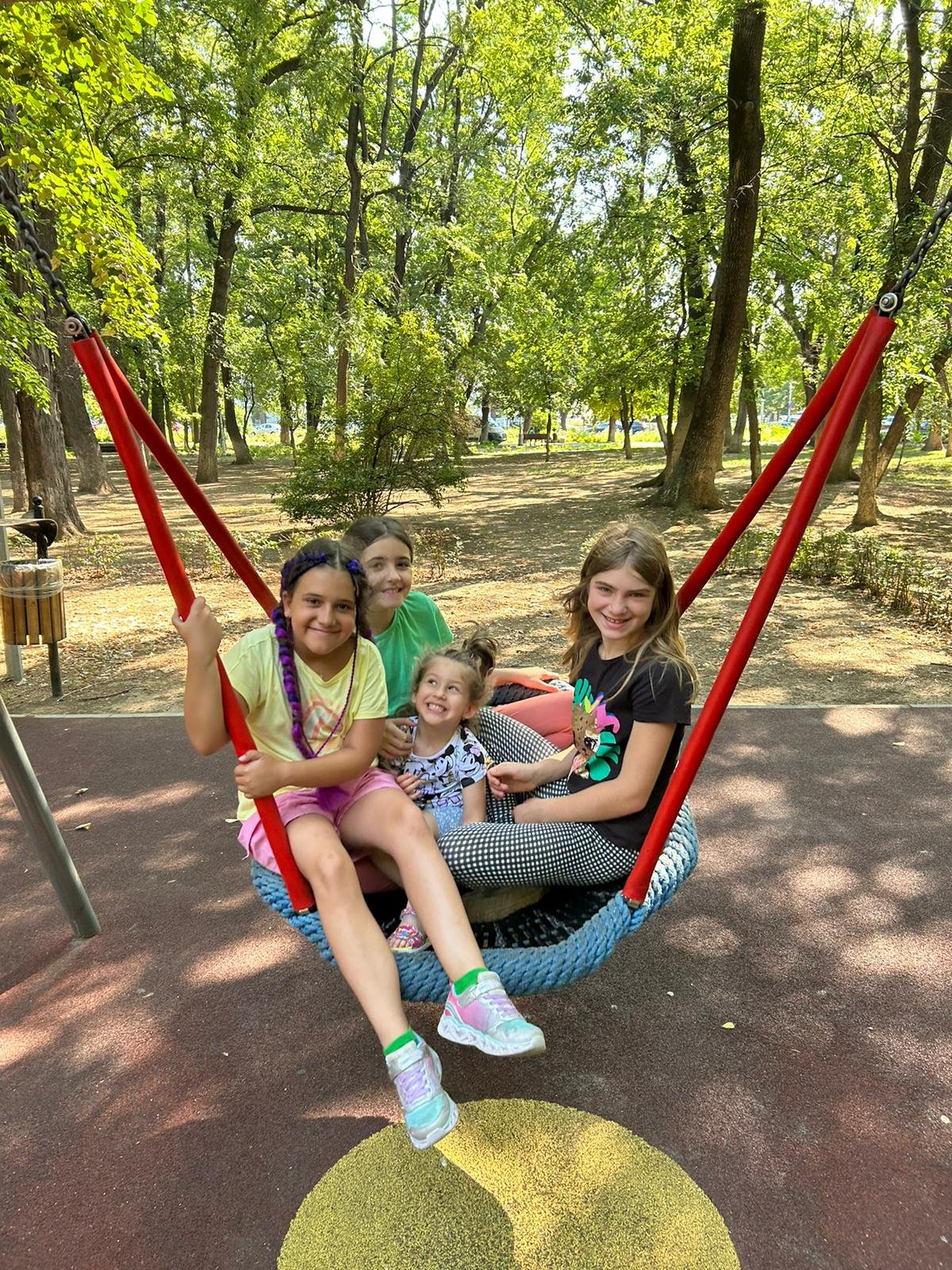 Four children sitting on a large swing in a park surrounded by trees.