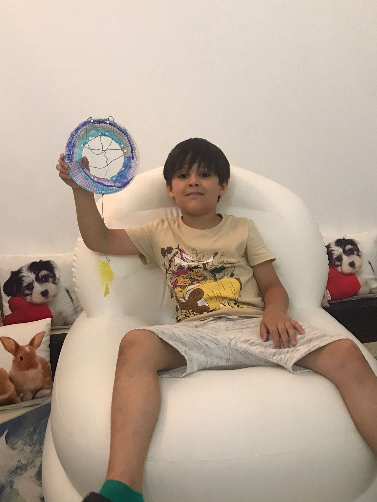 A young boy sitting on a white chair holding a colorful handmade dreamcatcher. There are pillows with images of dogs and a rabbit next to him.
