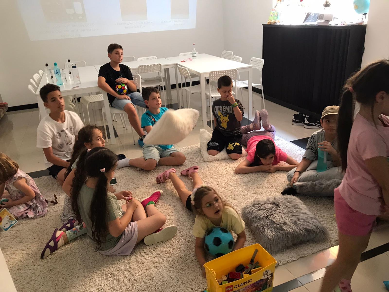 Kids sitting on a rug in a room, surrounded by pillows and toys, watching a projected screen. A yellow LEGO box is in the foreground, and a table with white chairs and water bottles is visible in the background.