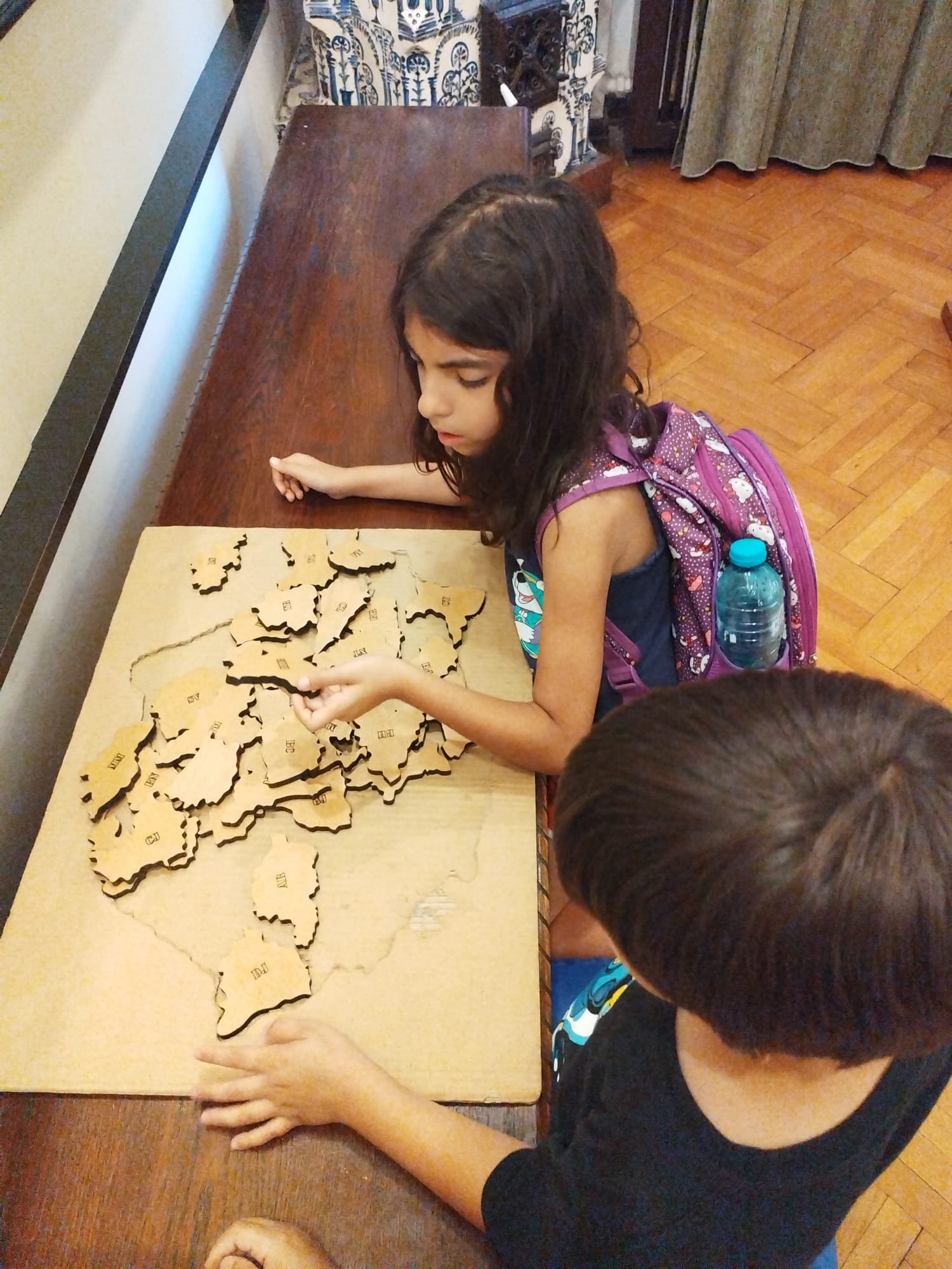 Two children playing with a wooden puzzle of a country's map on a table in a room with wooden flooring.