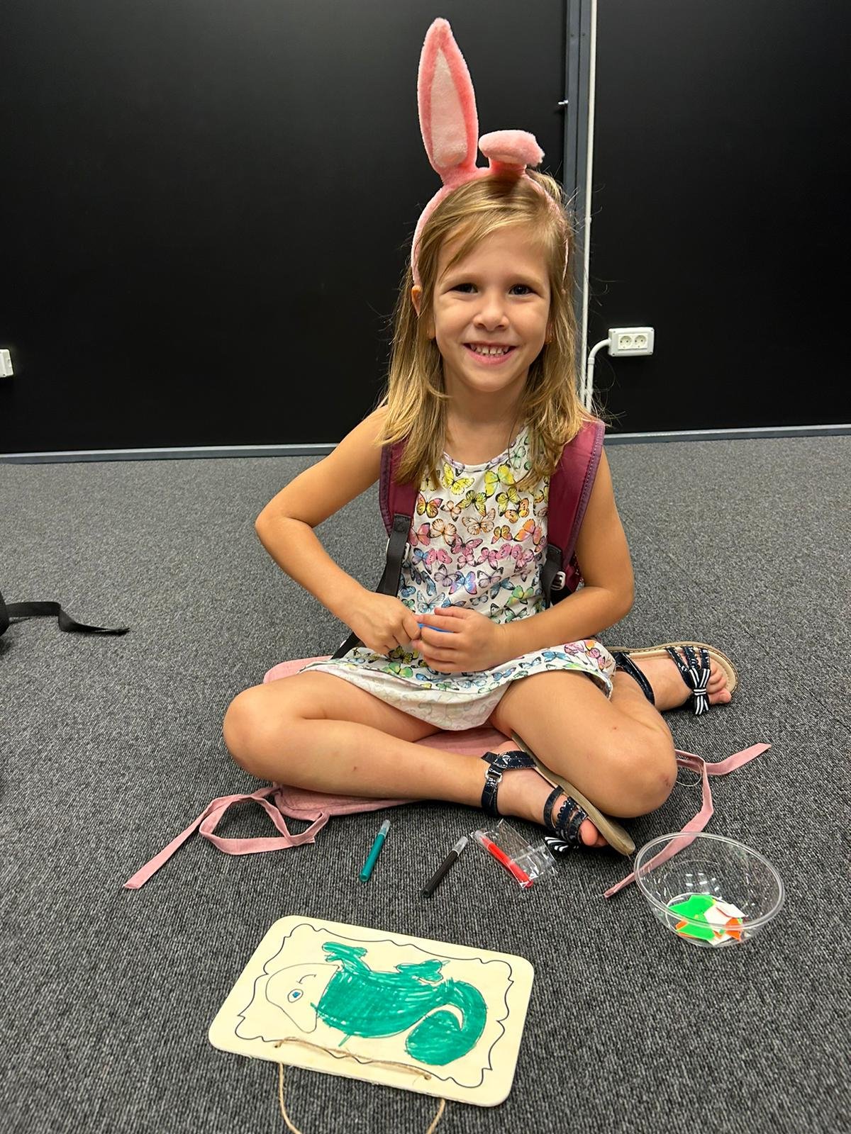 Smiling young girl wearing bunny ears and a printed dress, sitting on the floor with drawing materials and a colored artwork of a squirrel.