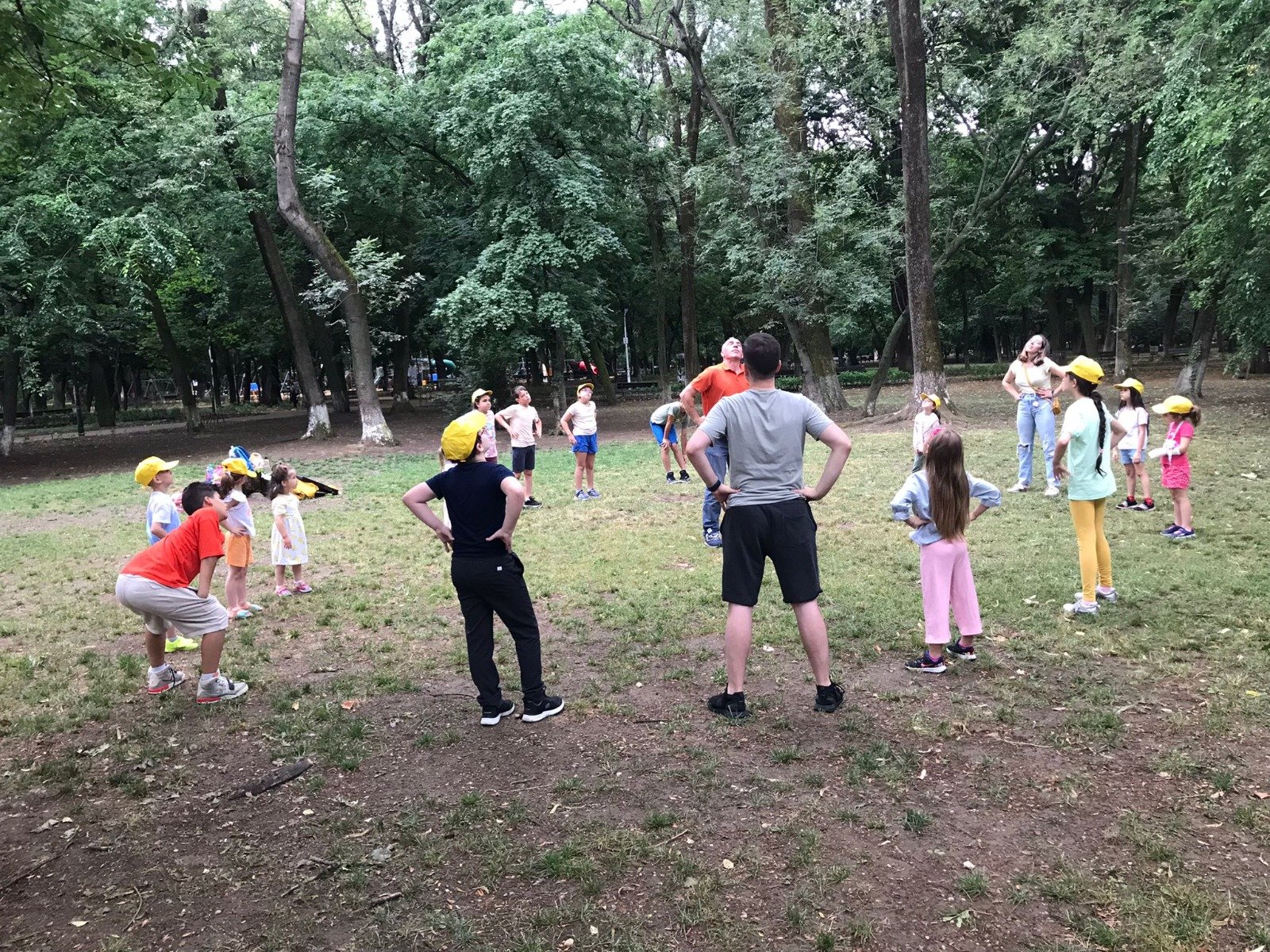 Group of children and adults playing a game in a park, surrounded by trees. The children, some wearing yellow caps, stand in a circle with adults participating in the center, engaging in an outdoor activity or exercise.