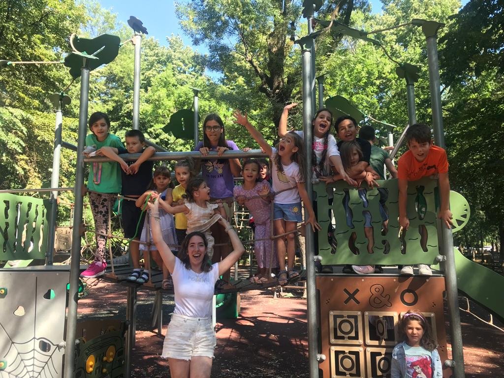 Group of children and an adult smiling on a playground structure with greenery in the background.