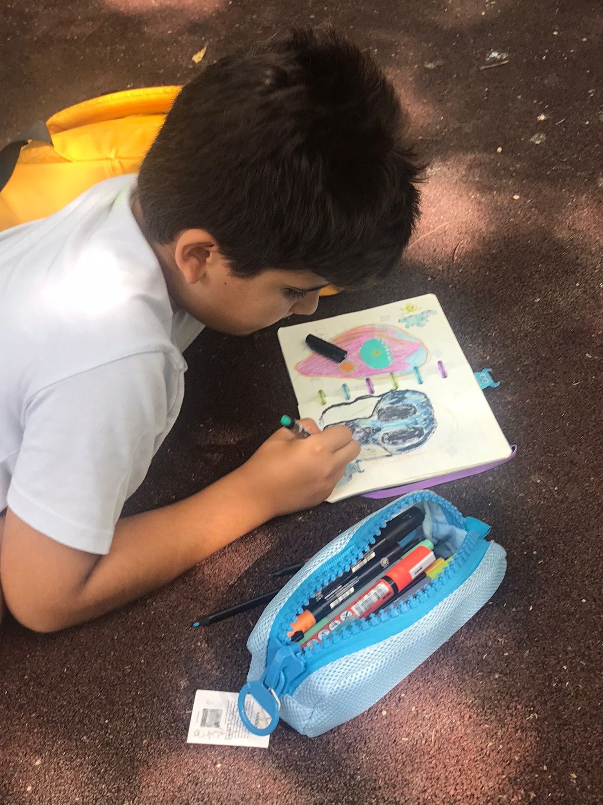 Child drawing in a sketchbook on the ground, using colored pens from a blue pencil case.