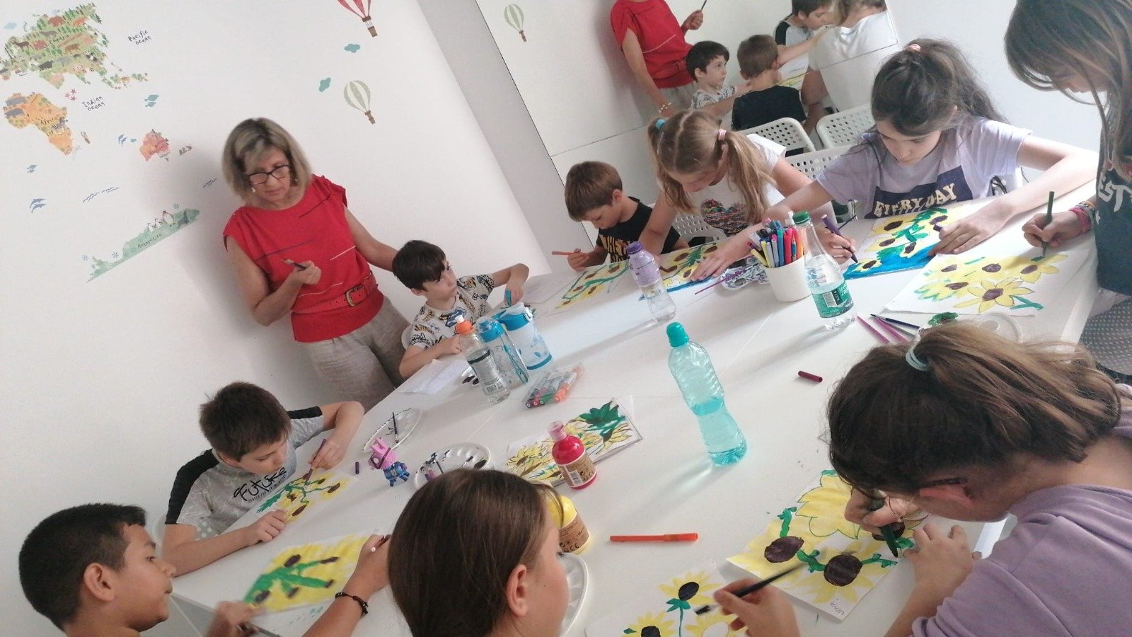 Children painting sunflower artworks at a table, supervised by a woman in a red top with a world map mural in the background.
