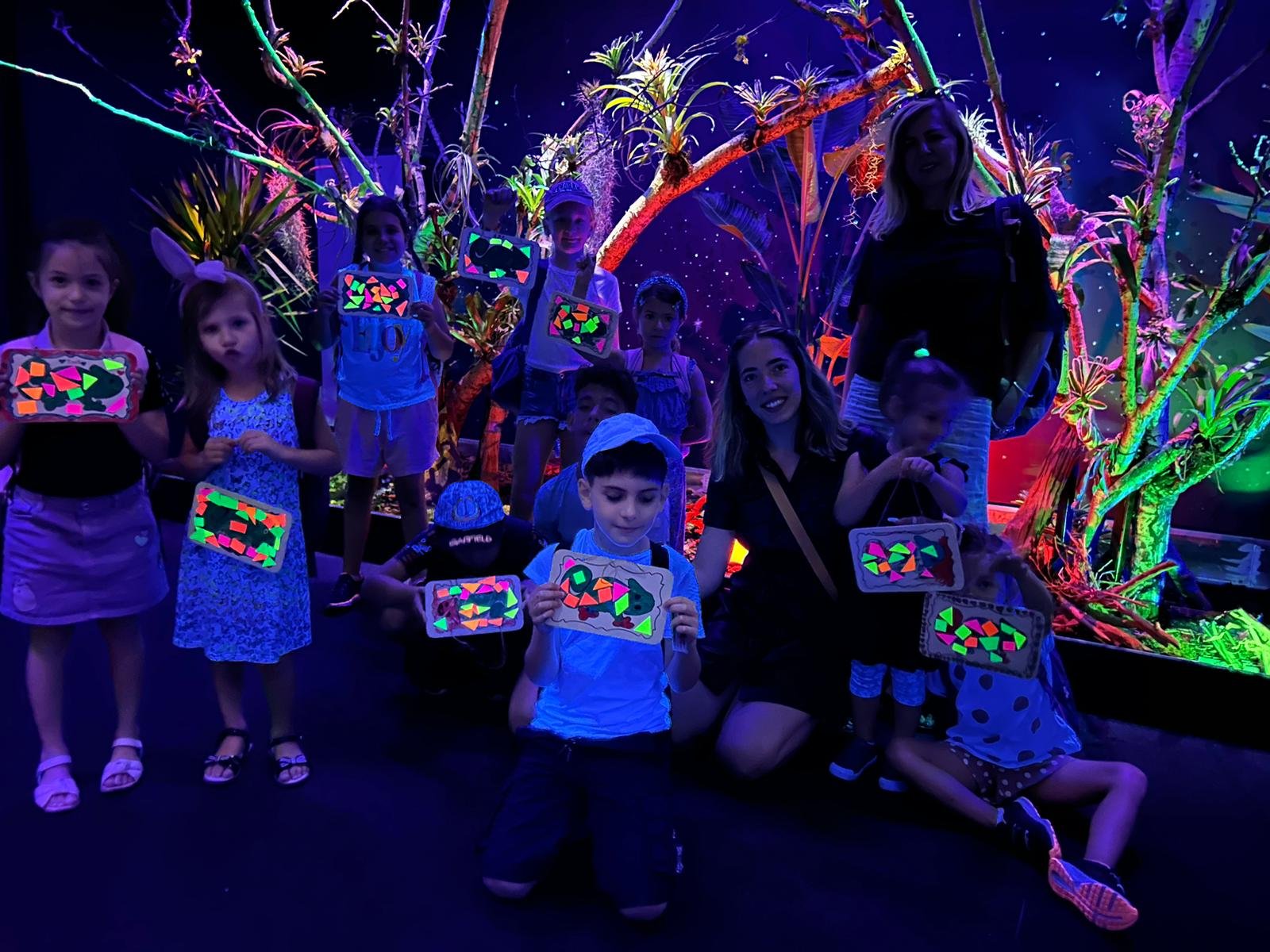 Children and adults posing with glow-in-the-dark artwork in a blacklight setting, surrounded by neon-painted plants and trees.