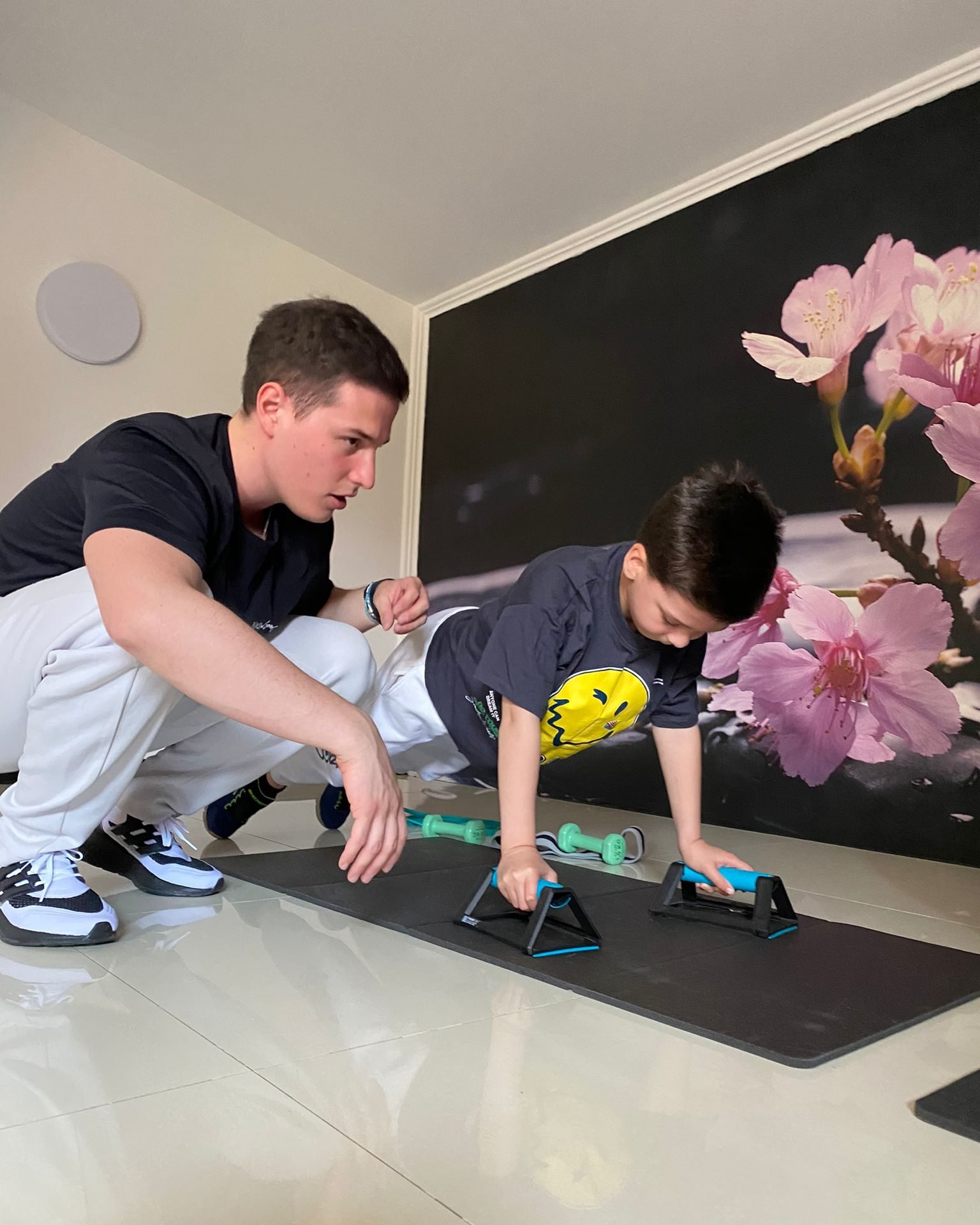 A child uses push-up bars on an exercise mat with adult supervision in a room with a floral wall mural.