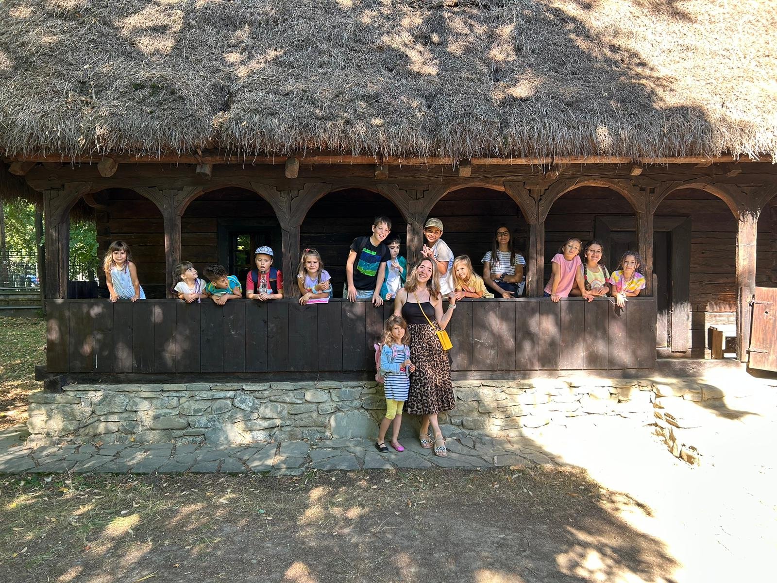 Group of children and adults on porch of rustic wooden house with thatched roof in sunlight.