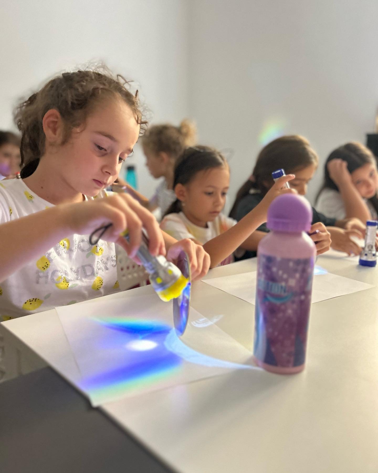 Children in a classroom using a prism to create light patterns on paper, with a colorful bottle on the table.
