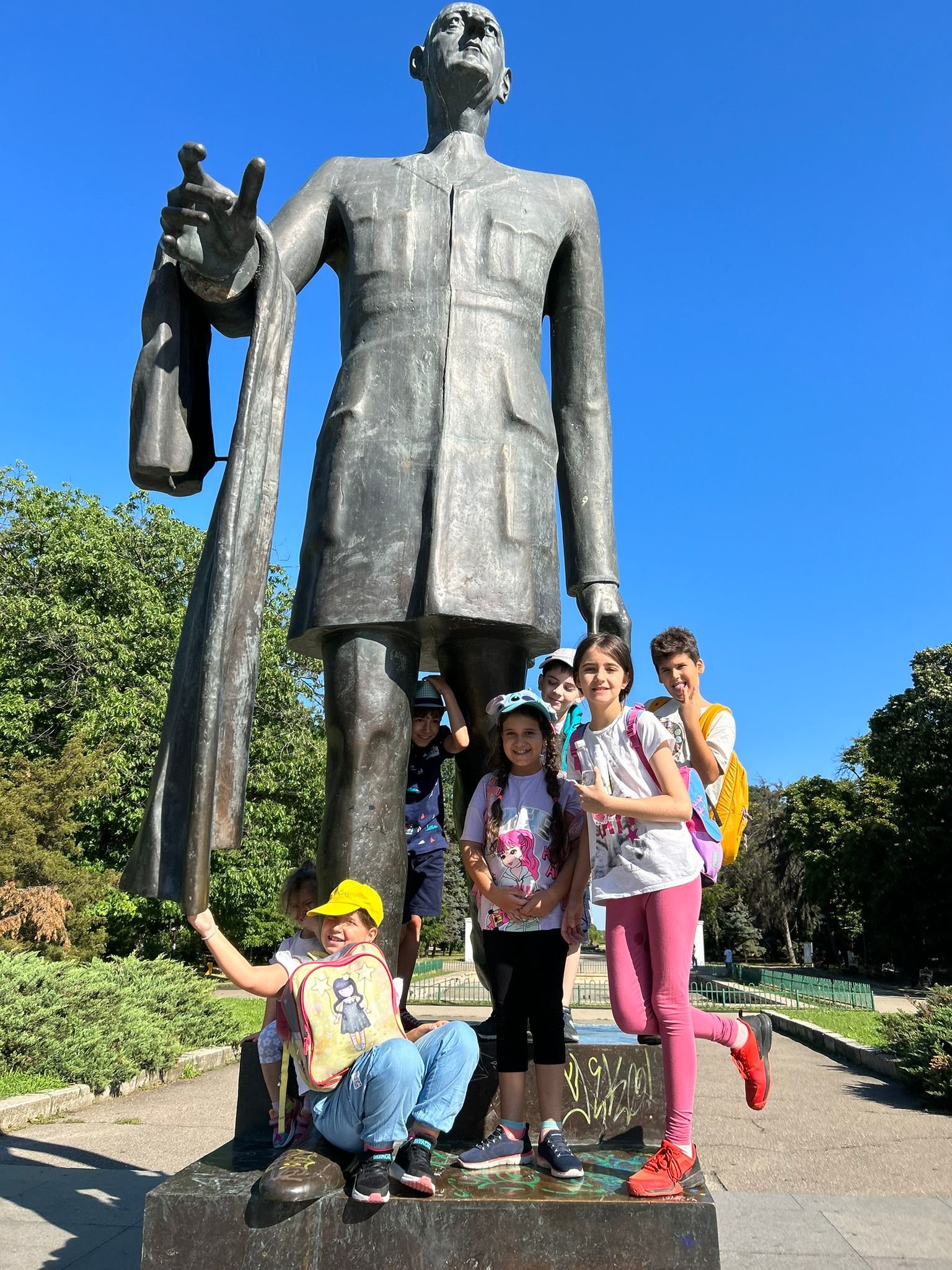 Group of children posing in front of a large statue in a park, with trees and a clear blue sky in the background.
