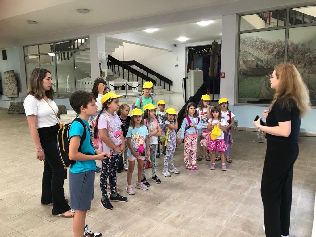 A group of children on a field trip, wearing yellow hats, standing in a museum-like setting, listening to a woman with long hair who appears to be guiding or instructing them. An adult woman is also observing the scene. The group is near a staircase