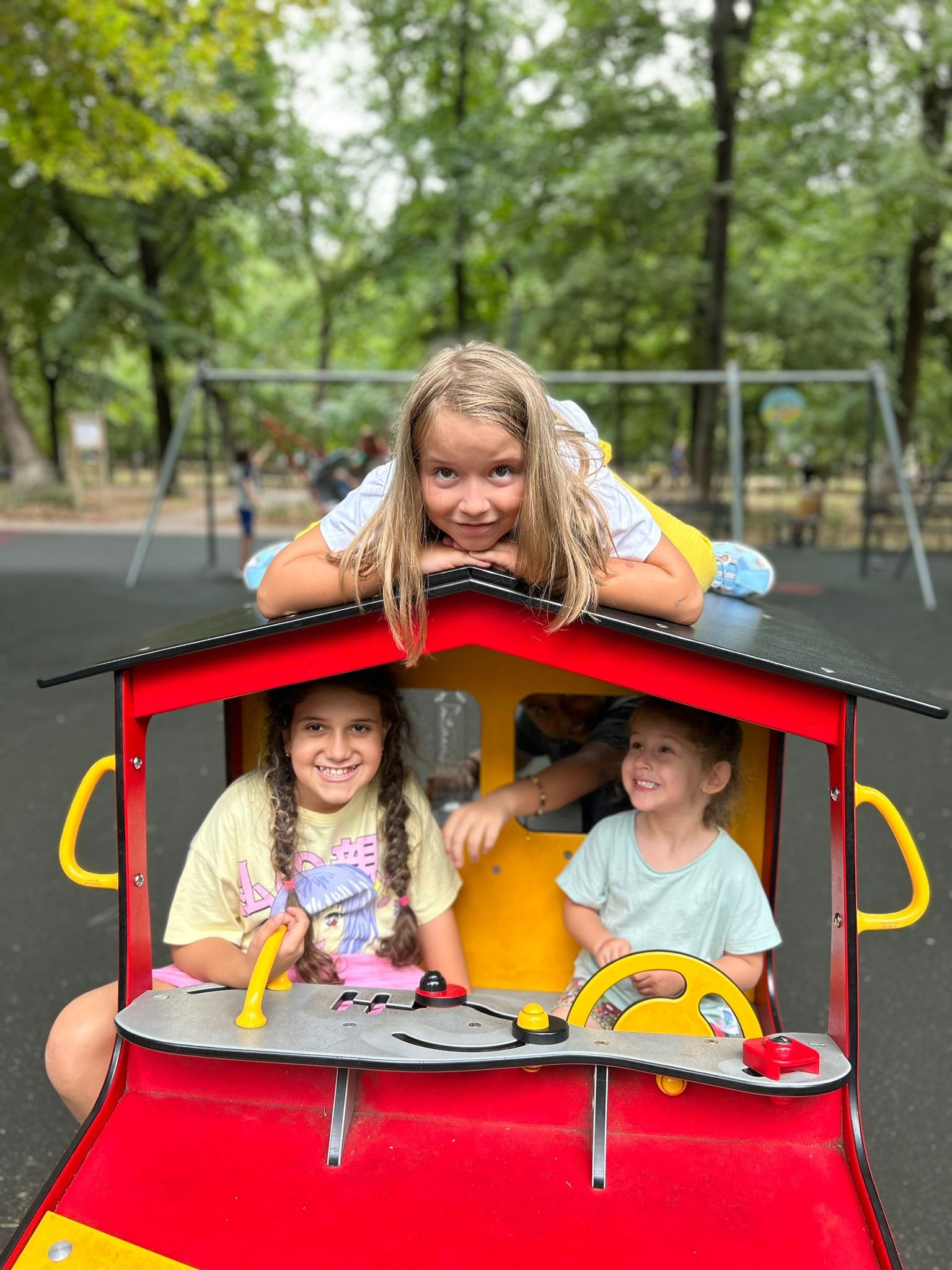 Children playing in a red play train at a park with green trees in the background.