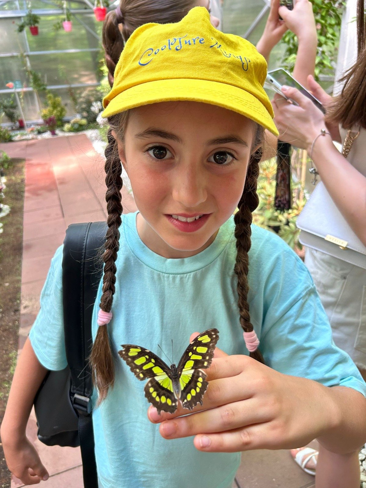 Child with a yellow hat holding a butterfly on their finger in a greenhouse.