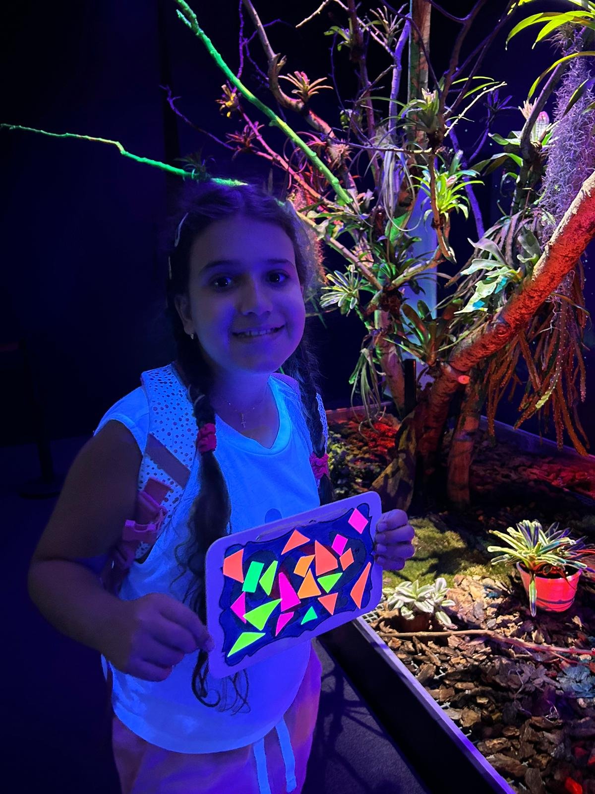 A young girl with long hair holds a blacklight-illuminated art piece with colorful geometric shapes. She stands in a dark room with glowing plants and branches.
