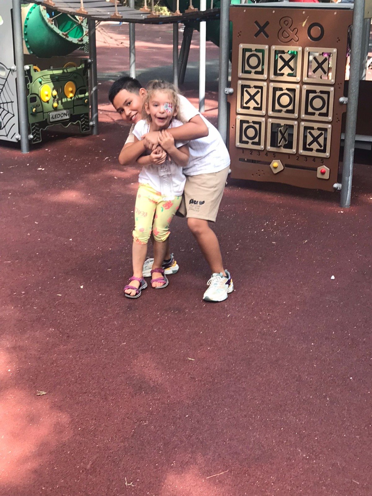 Two children playing on a playground, with one child hugging the other. There are play structures and a tic-tac-toe board in the background.
