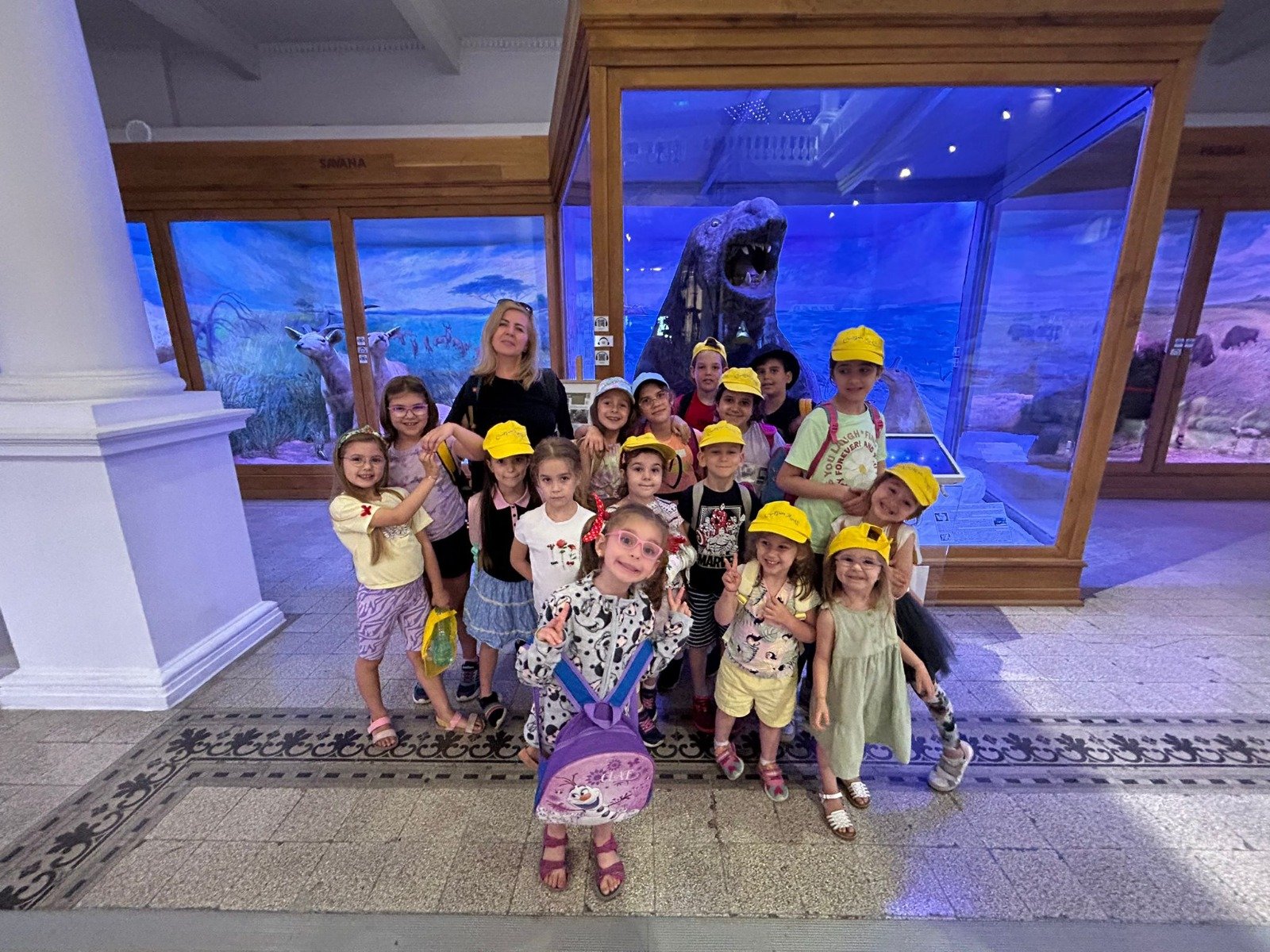 A group of children and an adult posing in a museum setting with a large animal exhibit in the background. The children are wearing yellow hats and backpacks.
