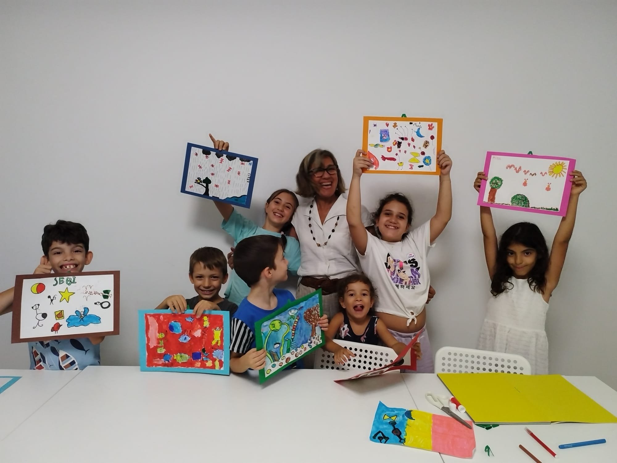 Children and an adult holding colorful drawings in a classroom setting, smiling and showcasing their artwork.