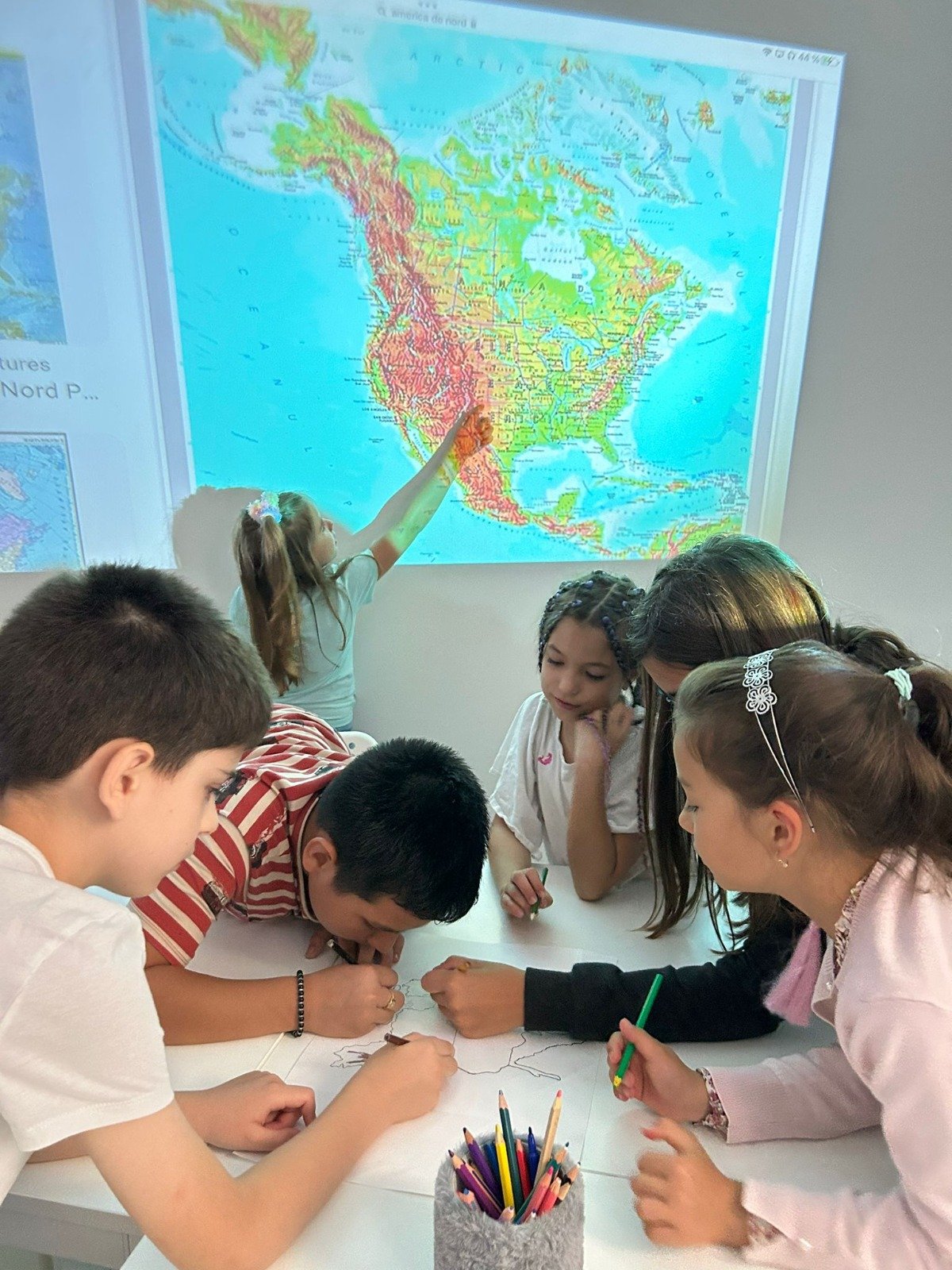 Children in a classroom working together on a map project, with a large map of North America projected on the wall. One child points at the map, while others draw on paper with colored pencils.