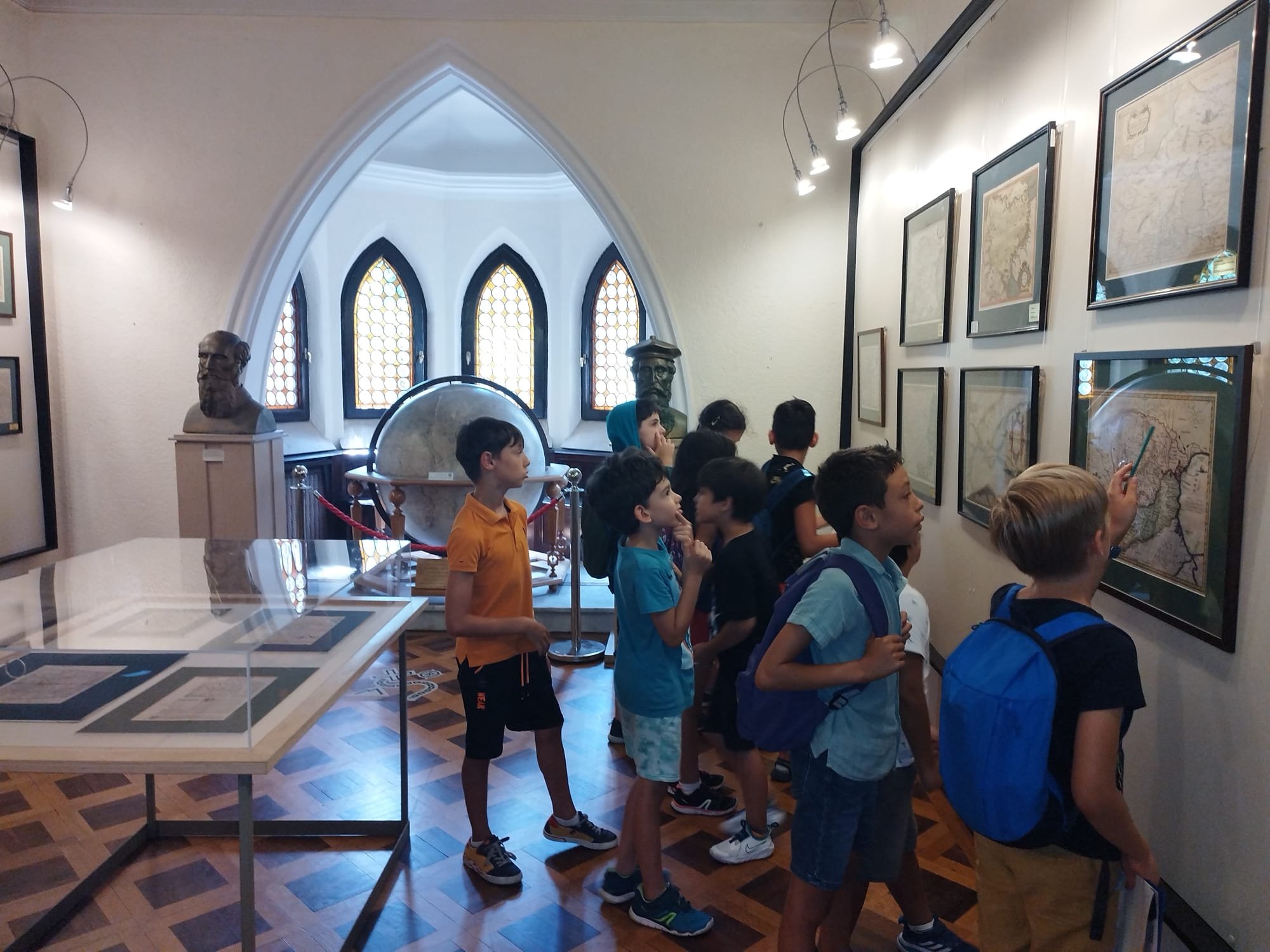 A group of children in a museum, observing framed maps on a wall. The room has geometric windows, wooden flooring, and displays a large globe and bust sculptures. A child points at a map with interest.