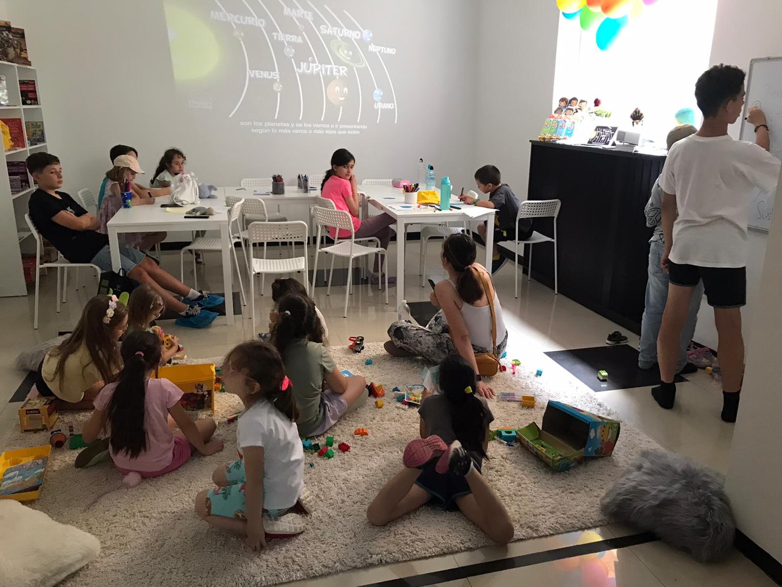 Children in a classroom sitting on chairs and on the floor, engaging in educational activities with a projection of the solar system on the wall.