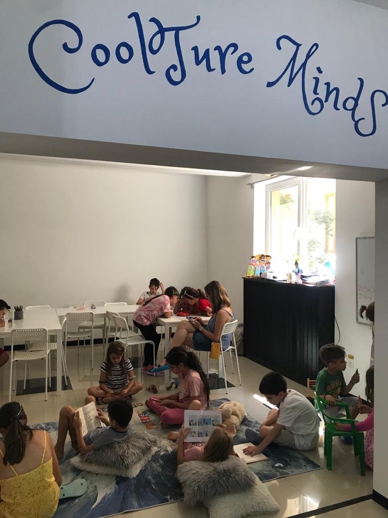 Children and adults in a reading room with "CoolTure Minds" sign, sitting at tables and on rugs with books.