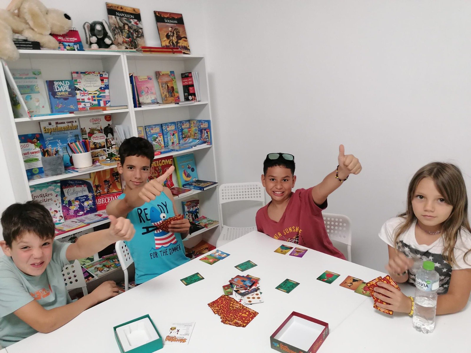 Four kids at a table playing a card game, surrounded by bookshelves filled with colorful books and stuffed toys.
