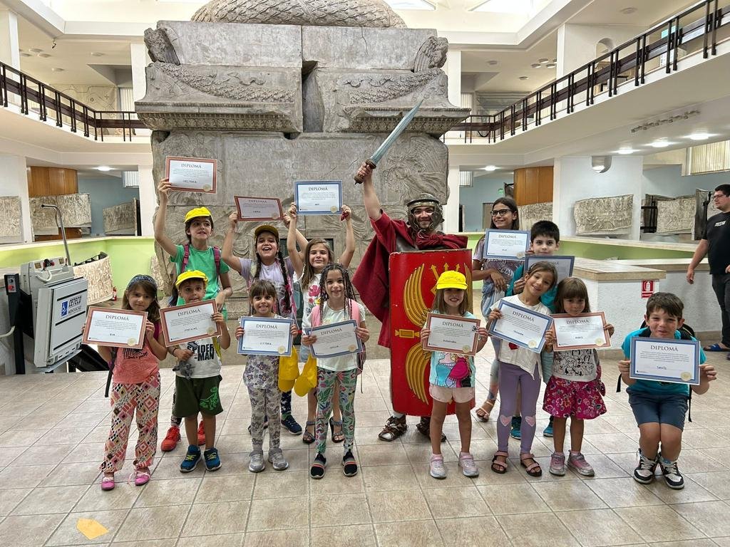 A group of children and an adult dressed as a Roman soldier holding diplomas or certificates standing in a museum-like environment with stone carvings in the background.
