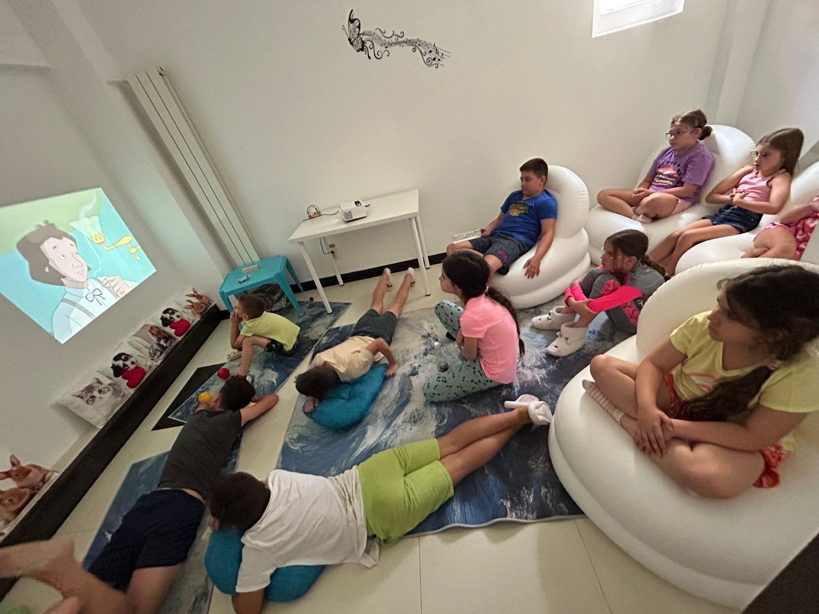 A group of children sitting and lying on the floor and bean bag chairs, watching a cartoon projected on a wall in a room. The room has cozy decorations including pillows and a small table.