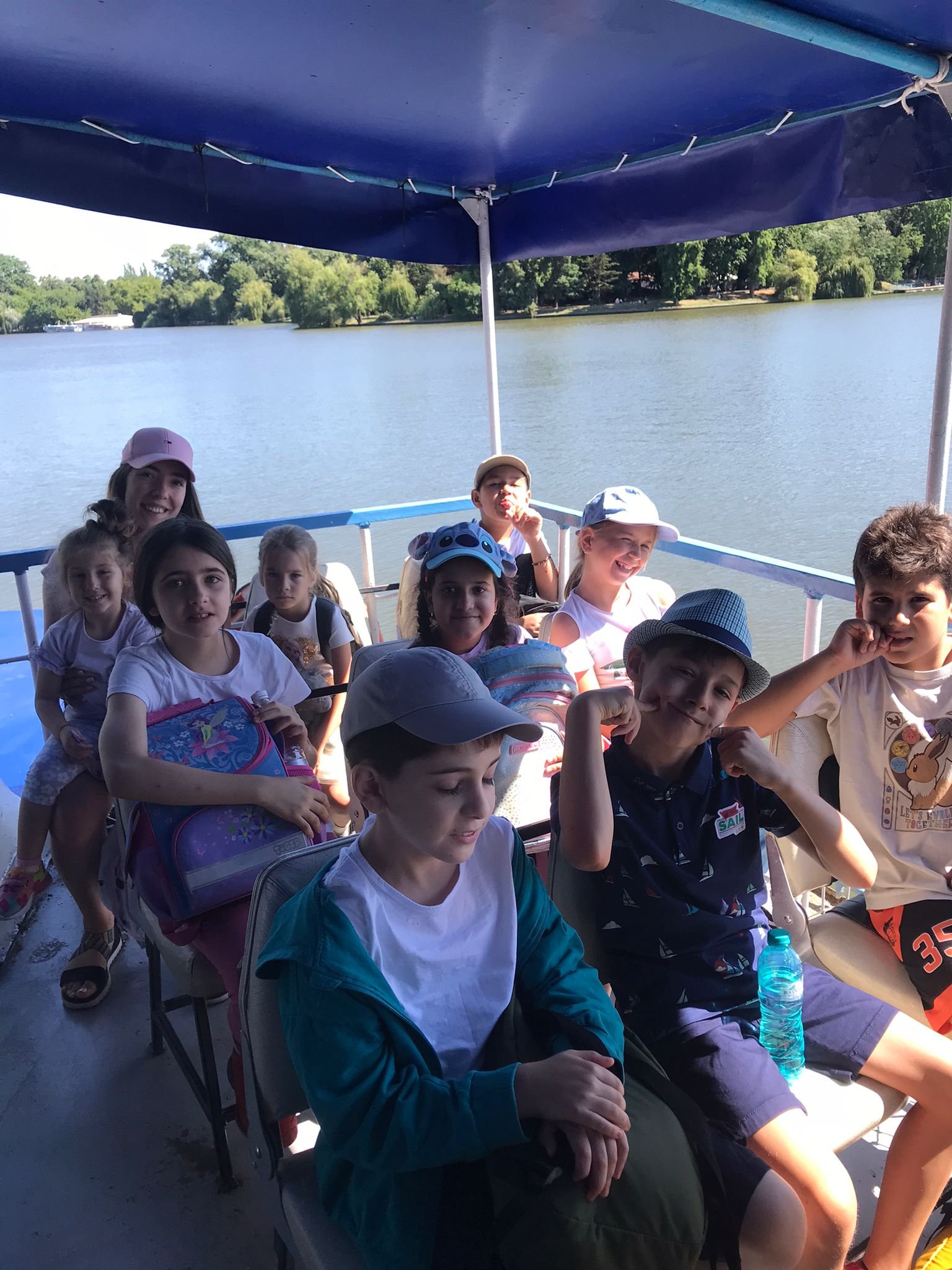 A group of children sitting on a boat under a blue canopy, with a lake and green trees in the background. They appear to be enjoying a boat ride on a sunny day. Some children are wearing caps and holding backpacks.