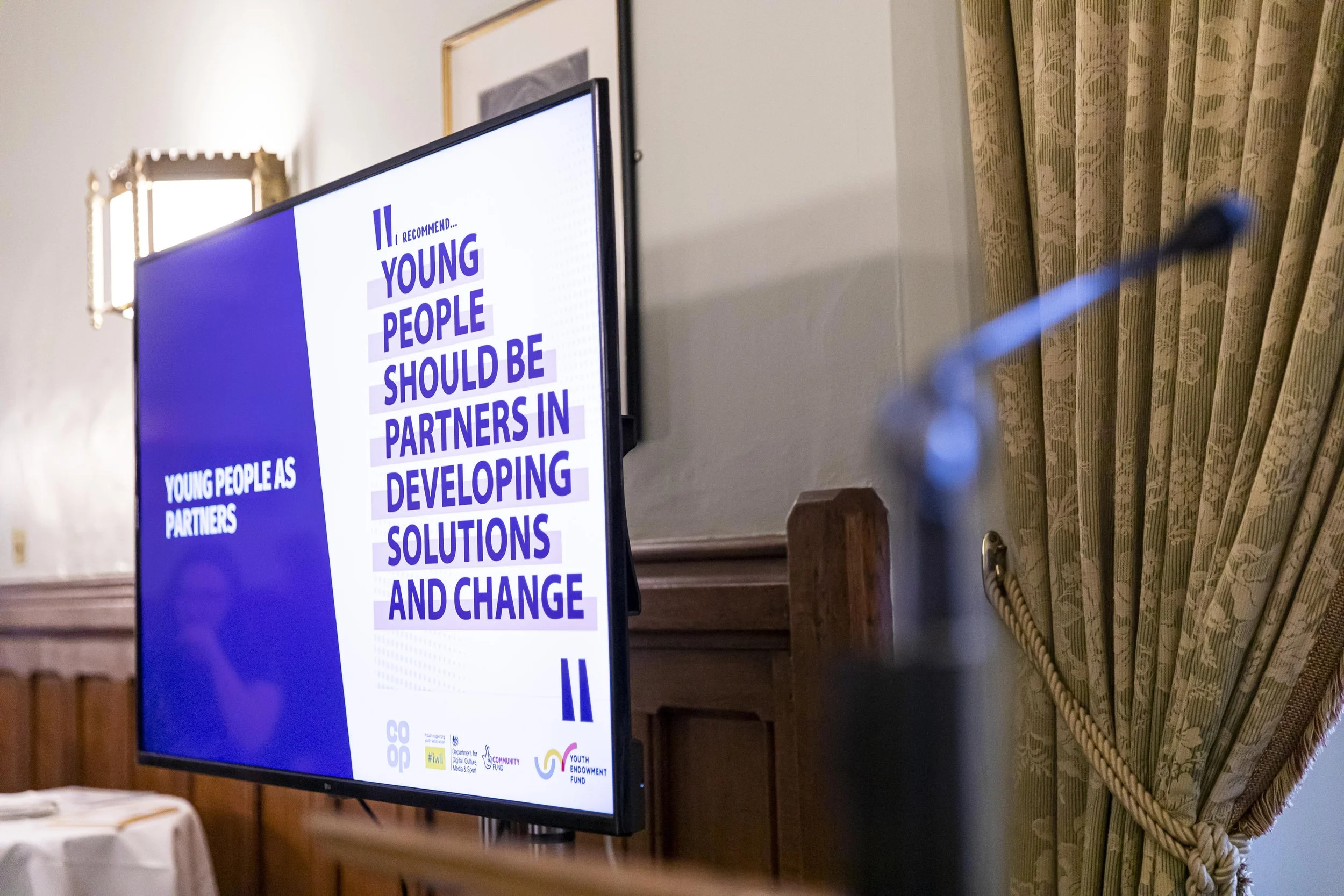 The image shows a television screen displaying a presentation slide with the text 'I recommend... Young people should be partners in developing solutions and change.' The background of the slide is mainly white and blue. The room has a wooden table, 