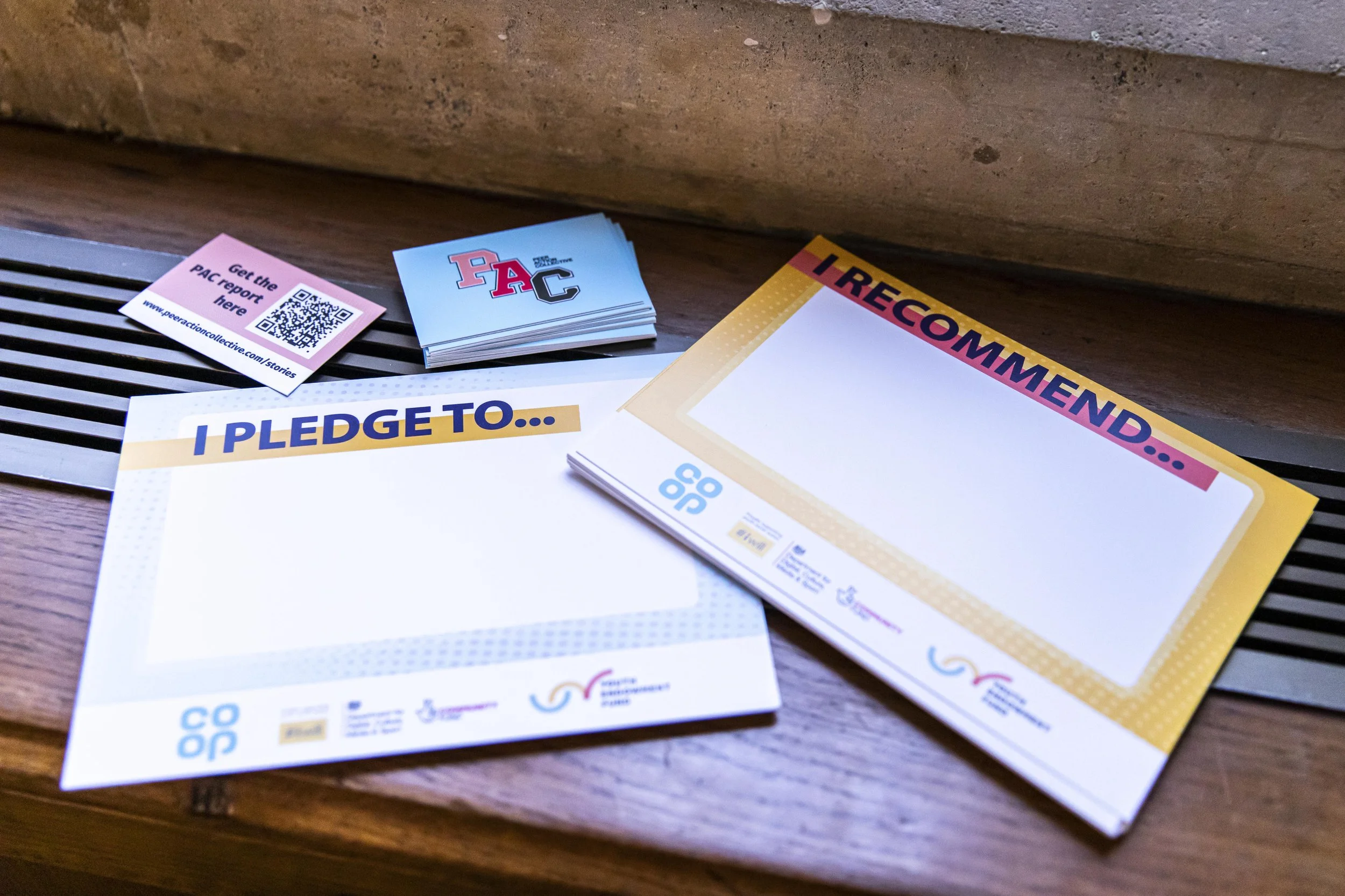 Brochures and flyers on a wooden surface, including a pink card about the PAC report, a stack of blue cards with autocross logos, and a yellow flyer with the words 'I RECOMMEND' visible.