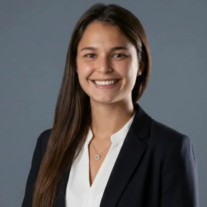 Hannah Herbst outdoors with long brown hair, smiling and wearing a navy blue shirt with a medical emblem and text, in a natural setting at sunset.