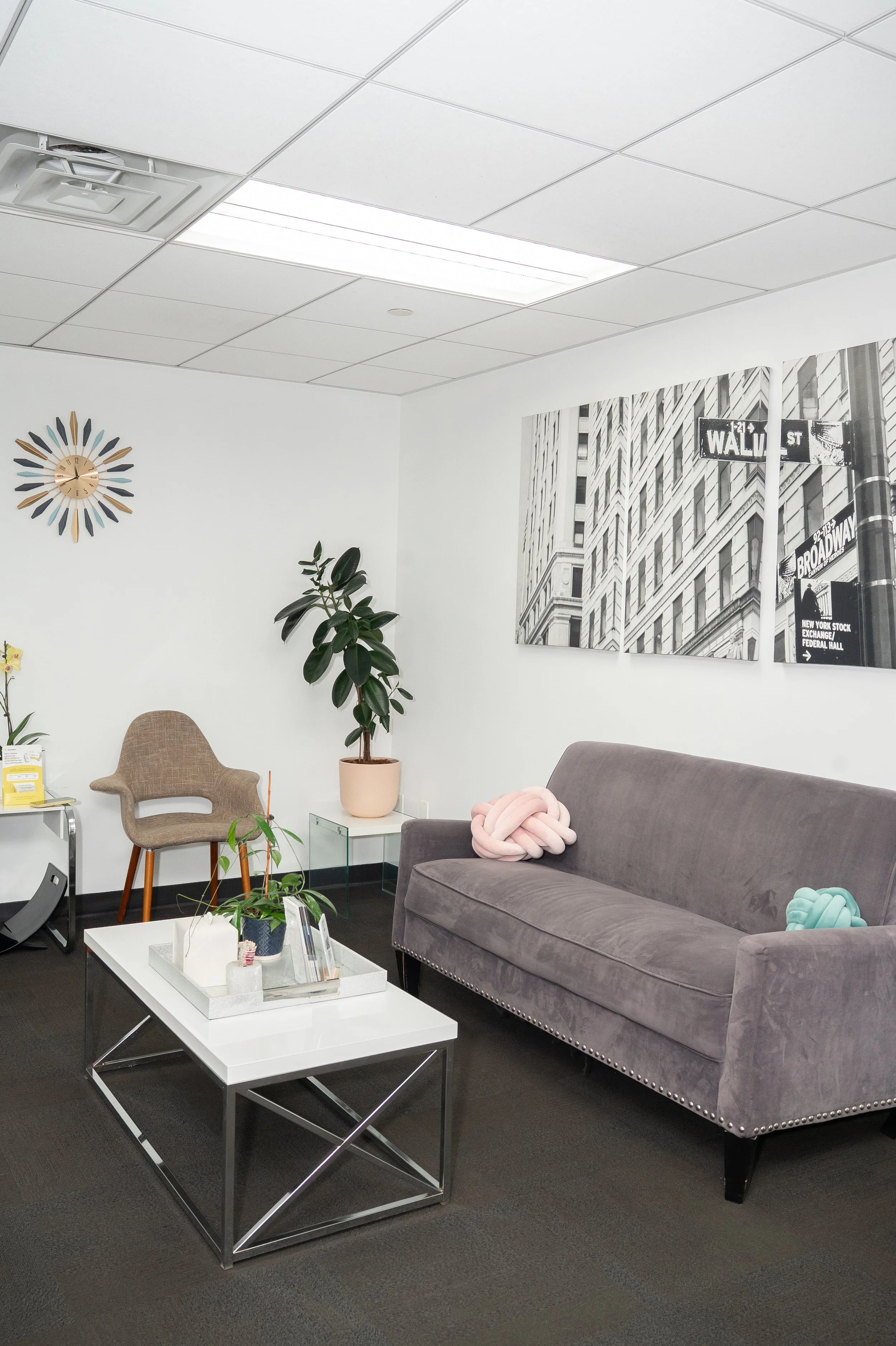 Modern office waiting area with a gray couch, armchair, coffee table, potted plants, wall clock, and a black and white Wall Street canvas print.